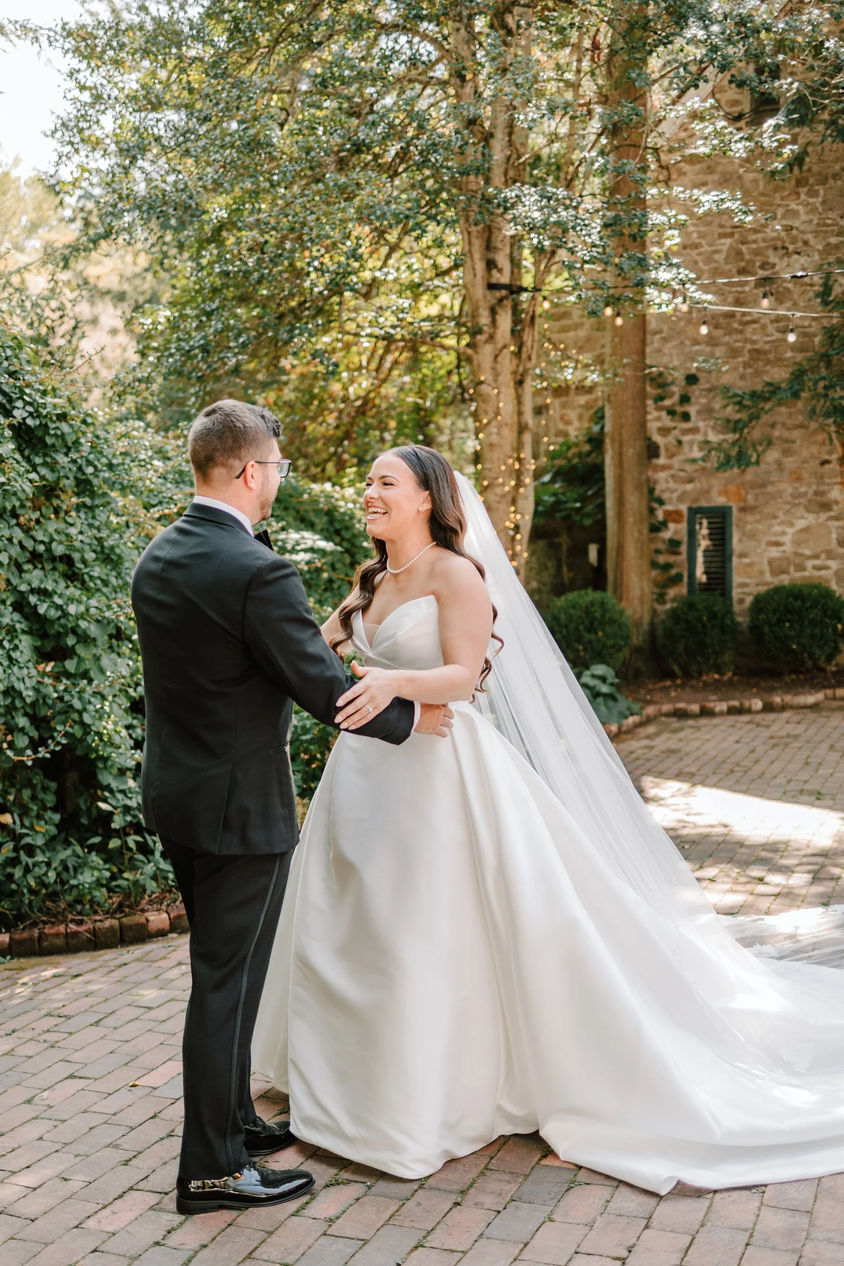 A bride and groom stand together outdoors, holding hands and smiling at each other during their wedding ceremony, with lush greenery and a stone wall in the background.