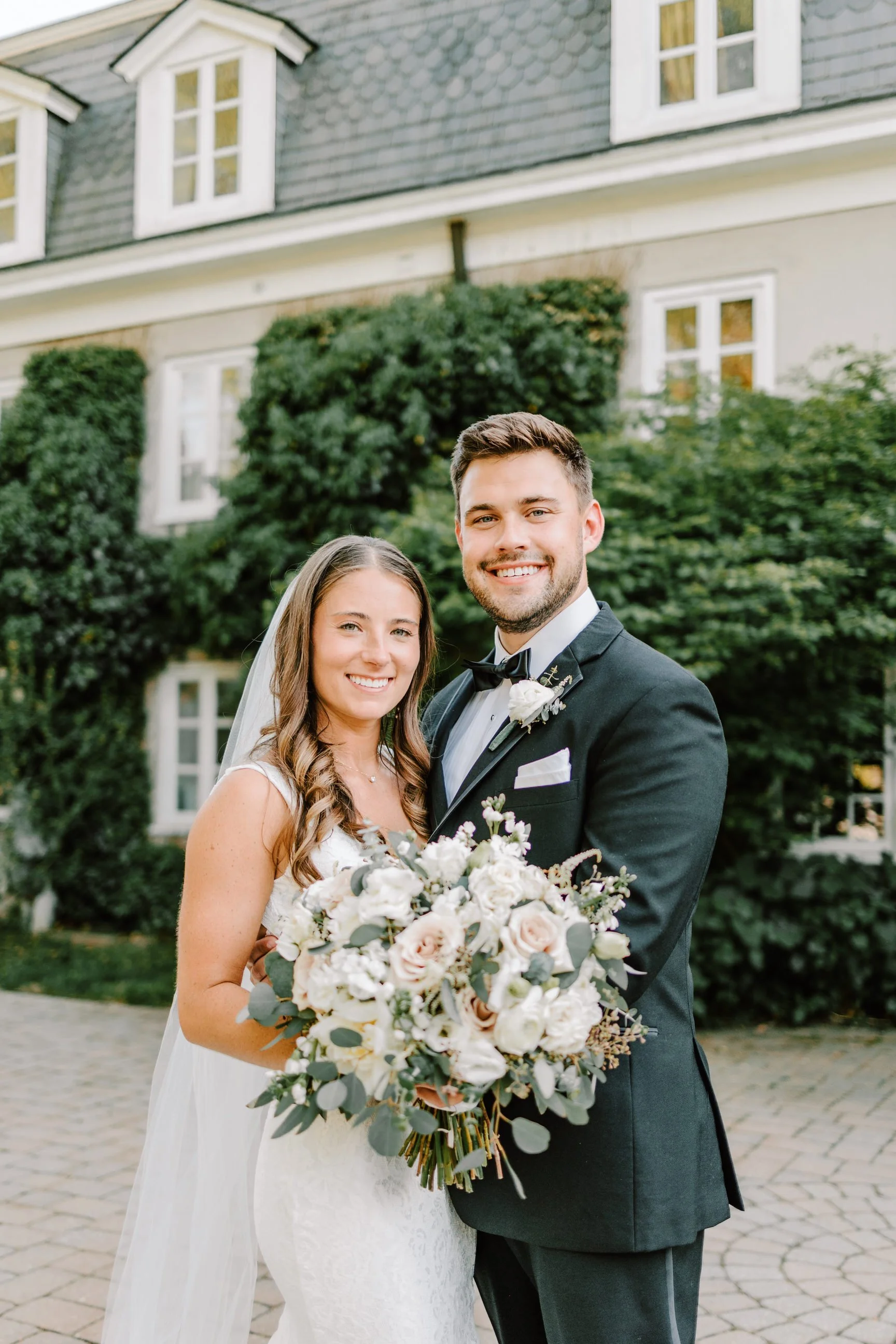 A newlywed couple standing outside in front of a house with greenery, smiling at the camera. The bride is holding a large bouquet of white and blush roses with greenery, and the groom is wearing a black tuxedo with a bow tie and boutonniere.