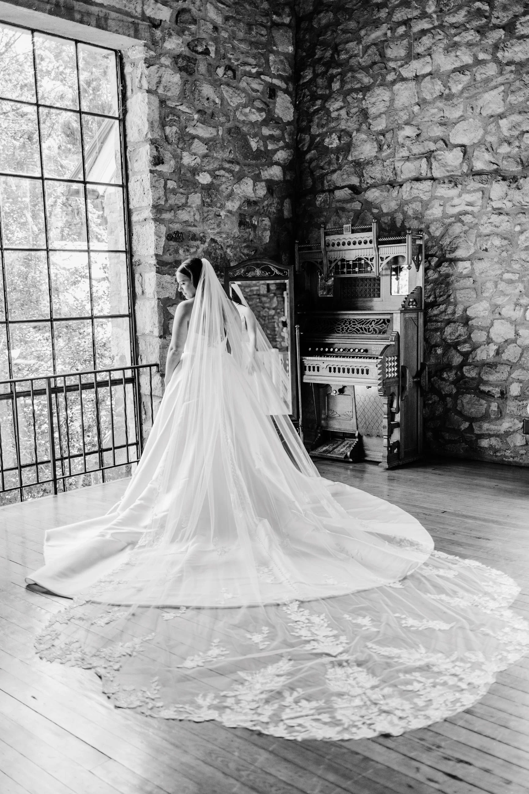 A bride in a wedding gown with a long train and veil standing on a wooden floor near a large window with natural light, a stone wall, and an antique wooden piano in a rustic interior.