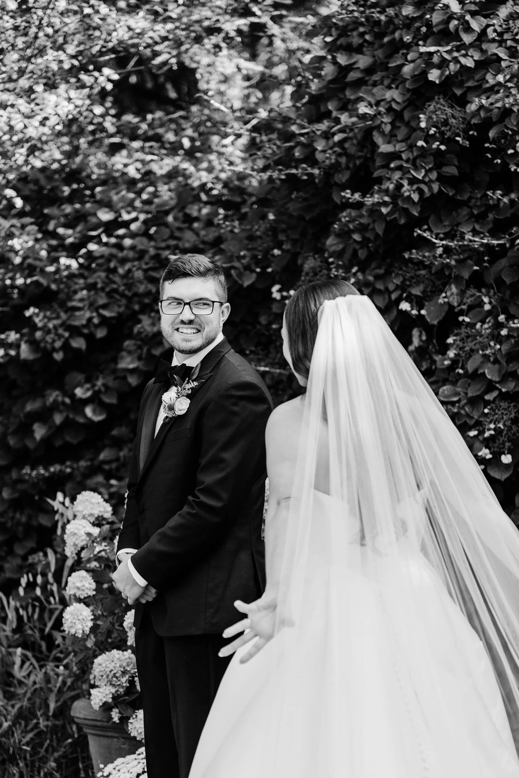 Black and white photo of a bride and groom outdoors, the groom with glasses in a tuxedo smiling at the bride, who is turned away from the camera, wearing a wedding dress and veil with a background of leafy bushes and flowers.