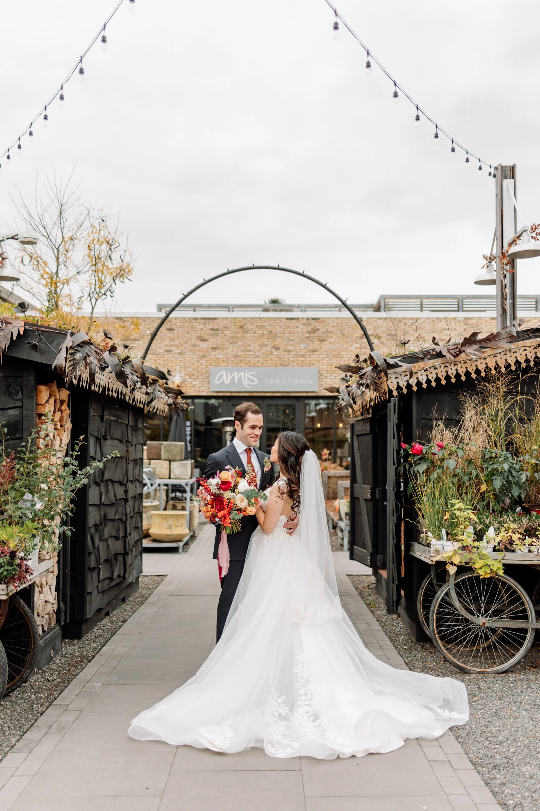 A bride and groom smiling at each other in an outdoor setting with string lights overhead and a bakery in the background.