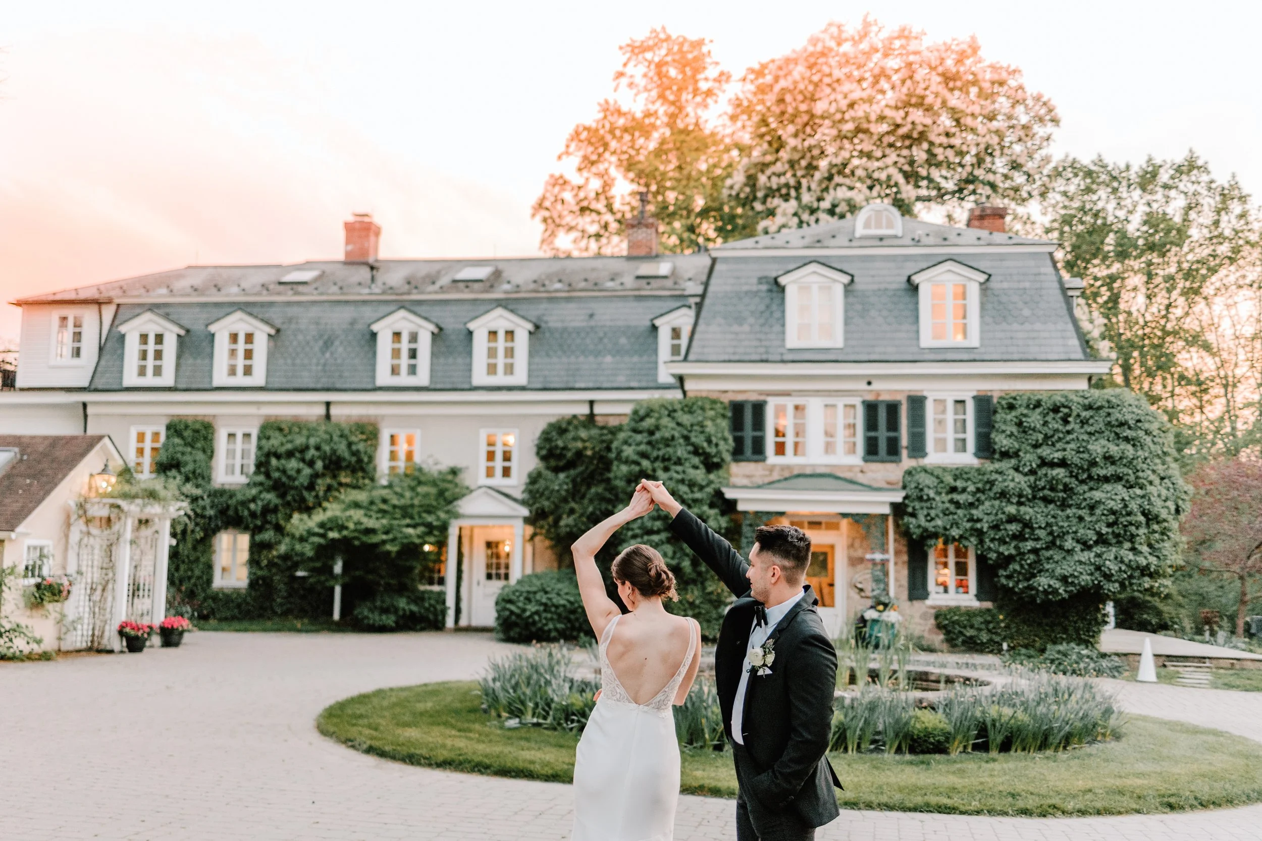 A wedding couple dancing outside a large, historic-looking house at sunset, with the bride in a sleeveless white gown and the groom in a black tuxedo. Philadelphia wedding photographer