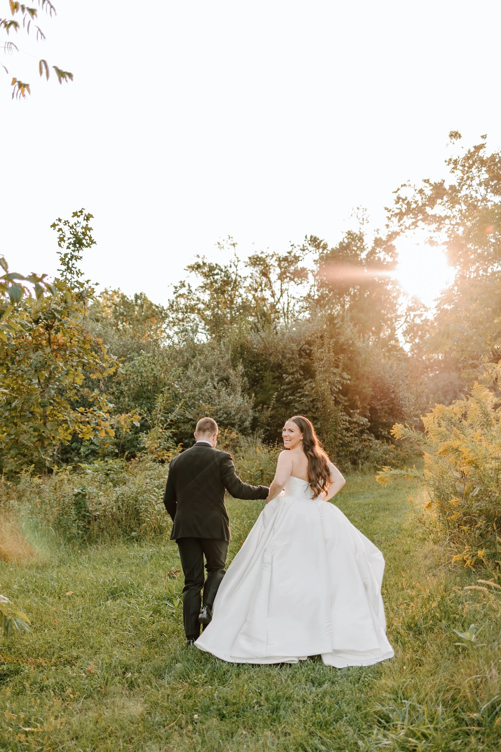 A bride and groom walking hand in hand on a grassy trail through a wooded area during sunset, with the bride in a white wedding gown and the groom in a black suit, both smiling.