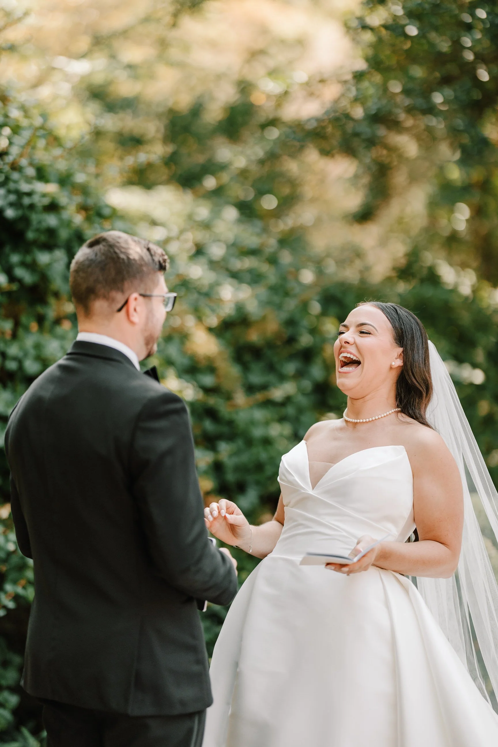 A bride and groom share a joyful moment during their wedding ceremony outdoors, surrounded by greenery.