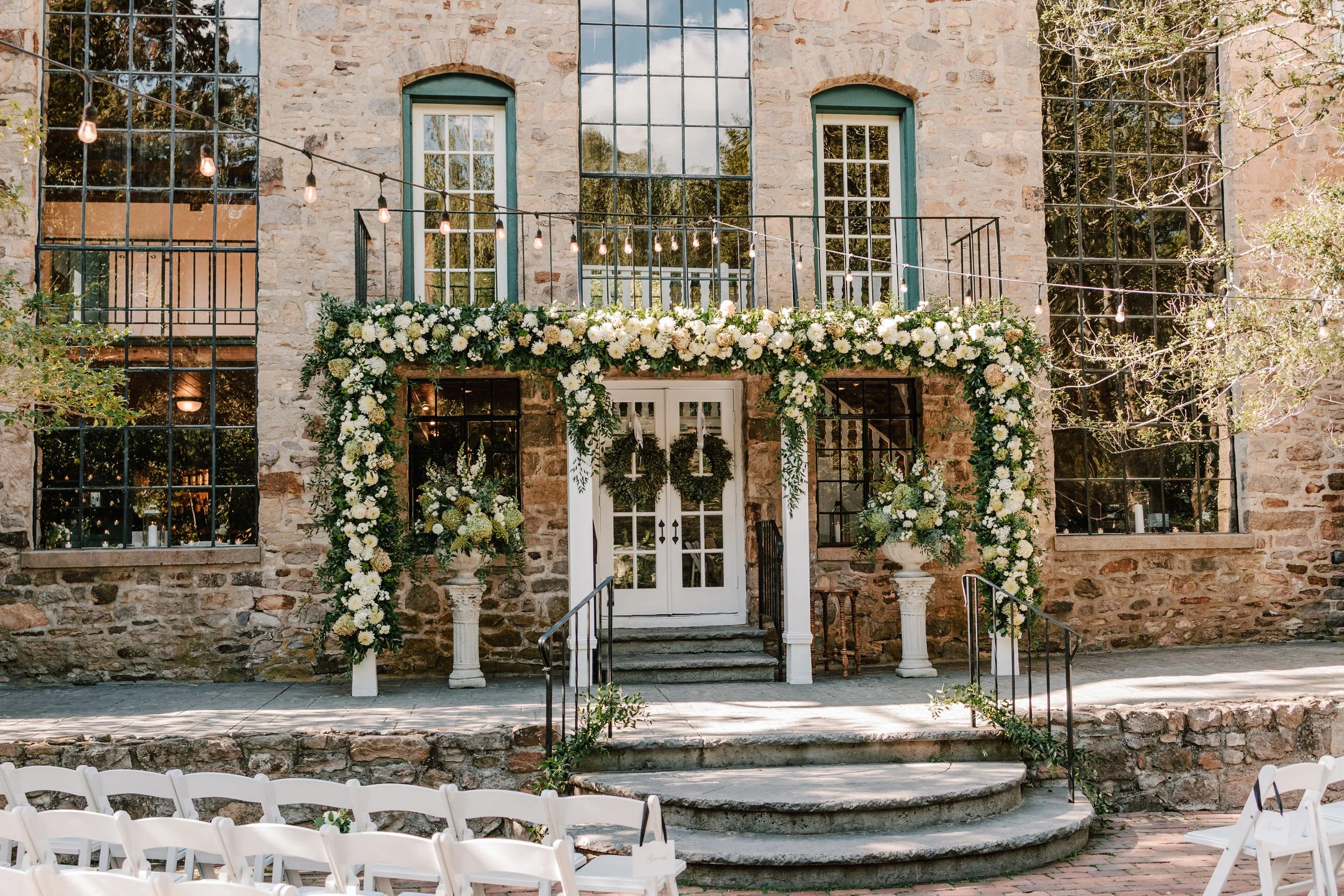 Outdoor wedding ceremony setup in front of a stone building with large windows. The entrance is decorated with white flowers and greenery, with some wreaths hanging on the door. There are white chairs arranged for guests, and string lights hang overh