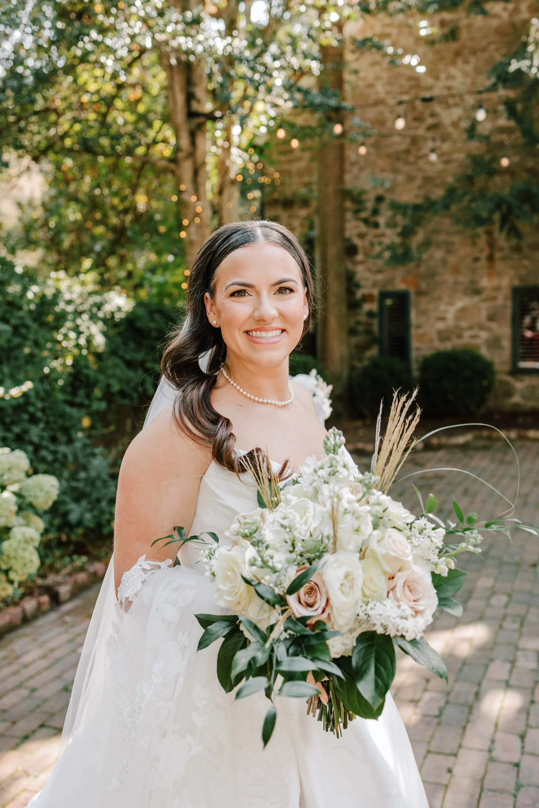 A smiling bride in a white wedding dress holding a bouquet of white and blush roses with greenery, standing outdoors in a garden with trees and string lights.