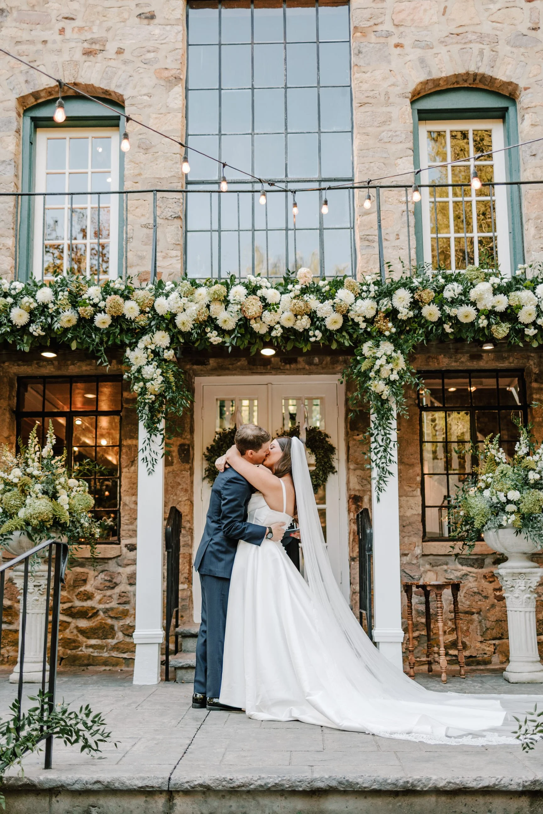 A bride and groom share a kiss on an outdoor wedding altar decorated with white flowers and greenery in front of a stone building with large windows and string lights.
