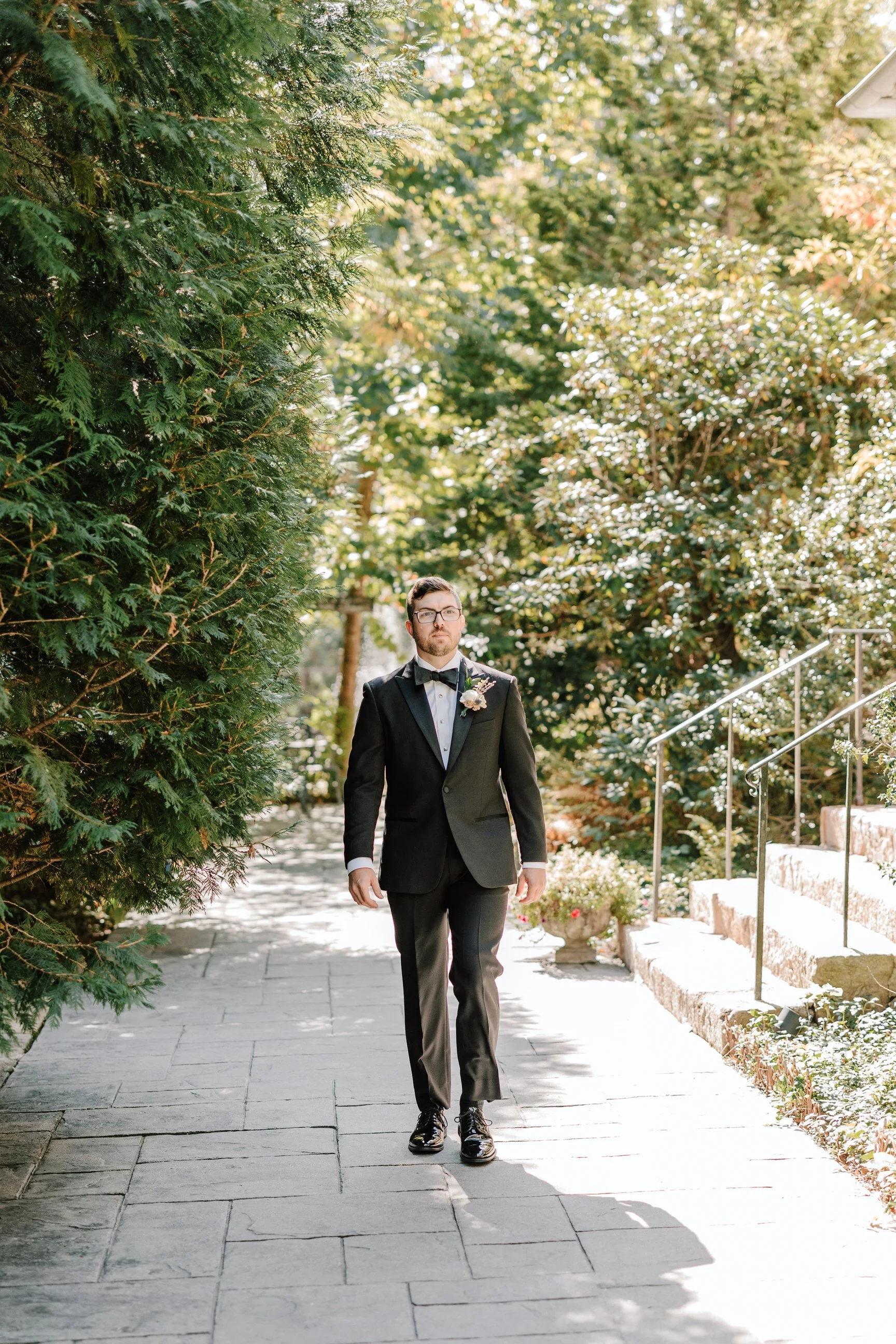 A man in a formal black tuxedo with a bow tie, walking outdoors on a stone pathway surrounded by lush green trees and plants.
