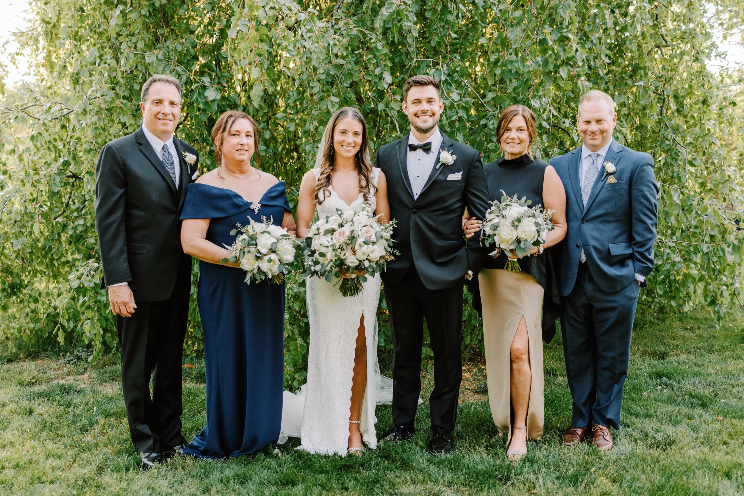 A wedding party standing outdoors in front of green trees, consisting of seven people dressed in formal attire, including the bride and groom in the center, surrounded by family members and friends holding bouquets of white and purple flowers.