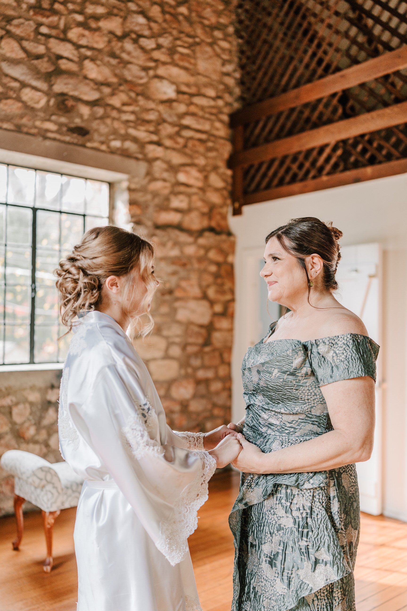 A bride and mother holding hands and smiling at each other inside a rustic room with a large window, exposed stone wall, and wooden beams. Holly Hedge Estate Sharyn Louise Photo