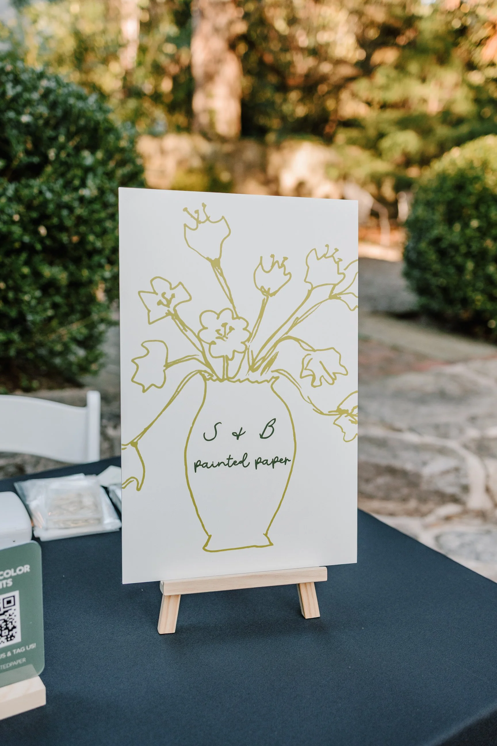 A white board with a simple yellow line drawing of a vase with flowers. The vase has the text 'S + B painted paper' written on it.
