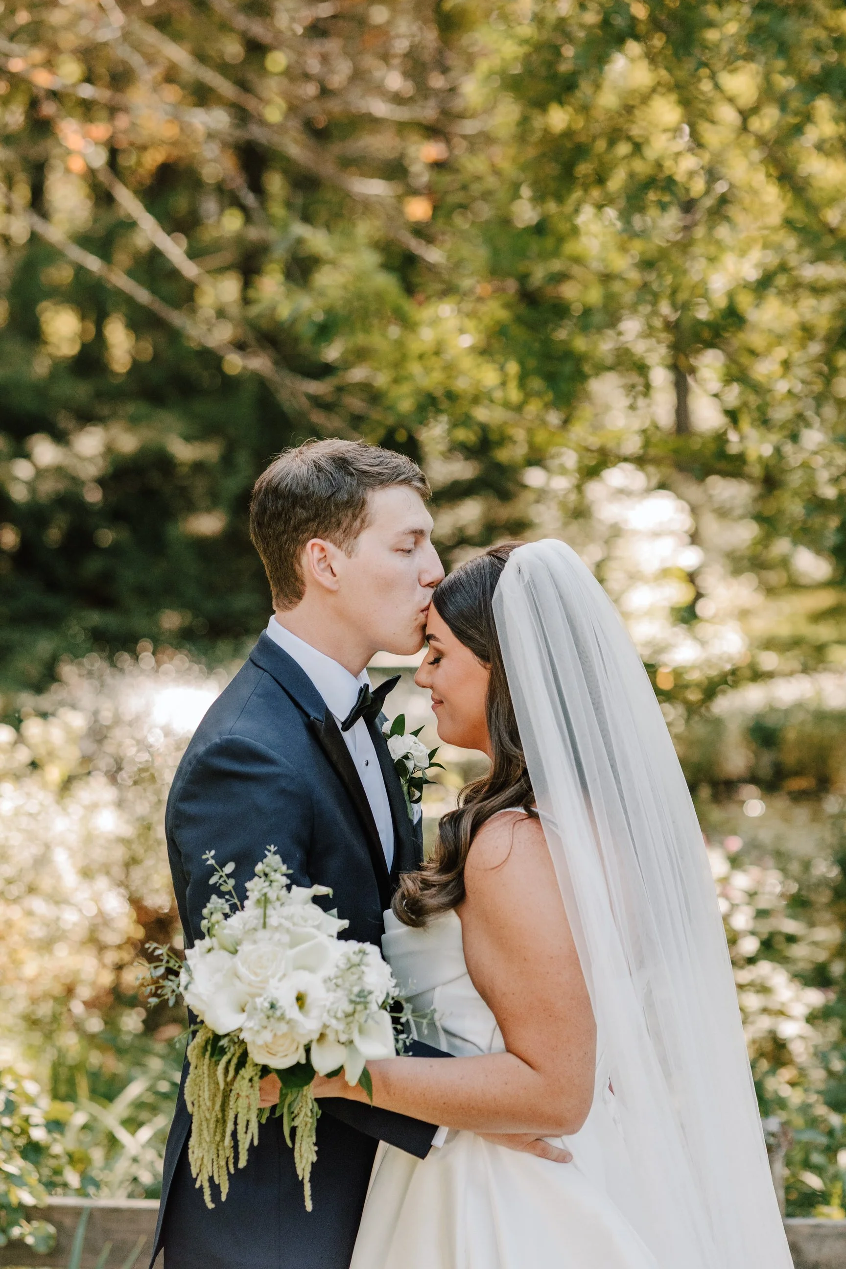 A bride and groom in wedding attire hugging outdoors surrounded by trees, with the groom kissing the bride's forehead and the bride holding a bouquet of white flowers.