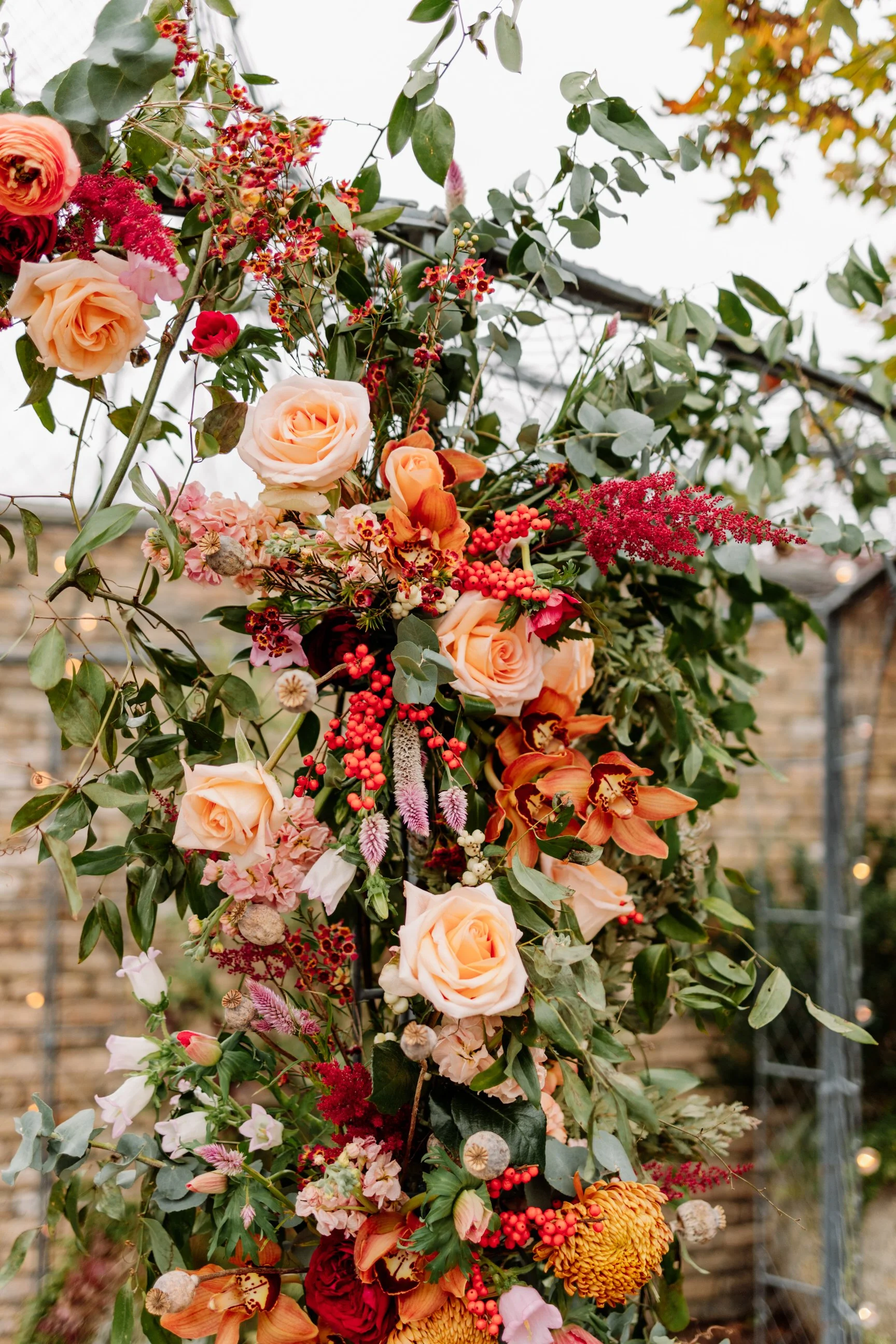 A floral arrangement with peach roses, orange orchids, red berries, pink and red flowers, and green leaves outdoors near a brick wall and a metal fence.