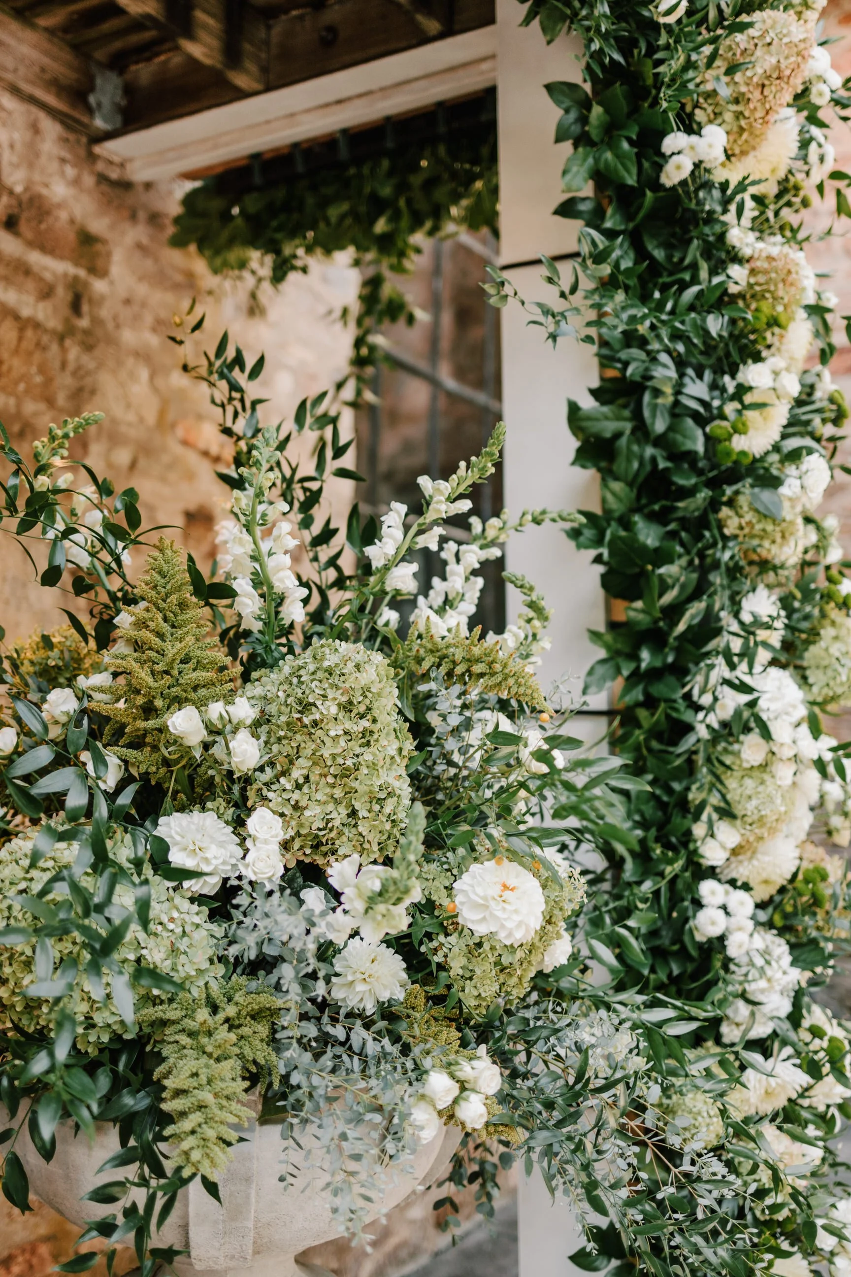 Close-up of a flower arrangement with white flowers, green foliage, and hydrangeas, set against a rustic brick wall and wooden structure.