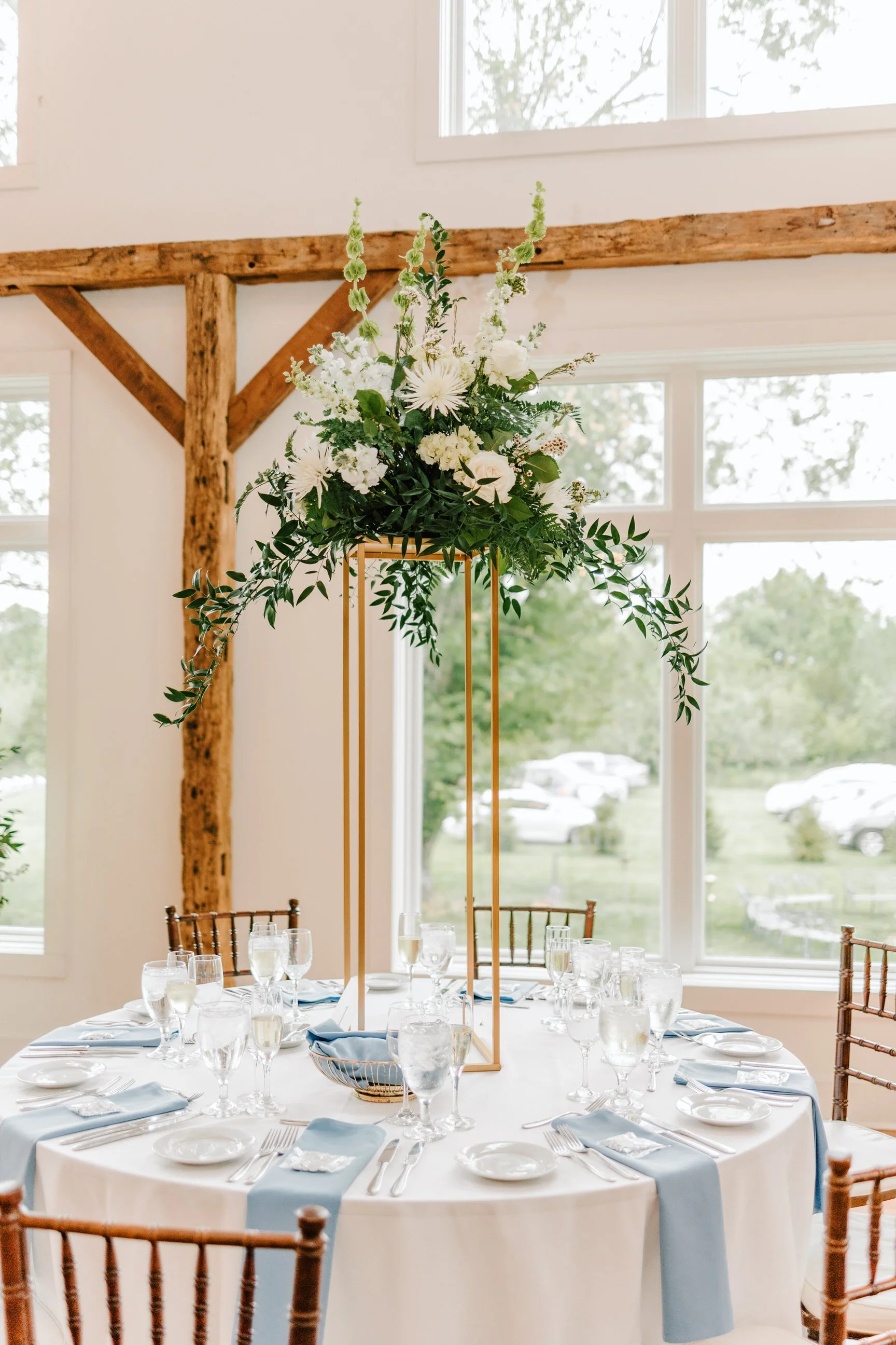 Round table set for a formal event with a floral centerpiece on a tall stand. The table has white tablecloths, blue napkins, and glassware. The setting is inside with large windows and wooden beams in the background.