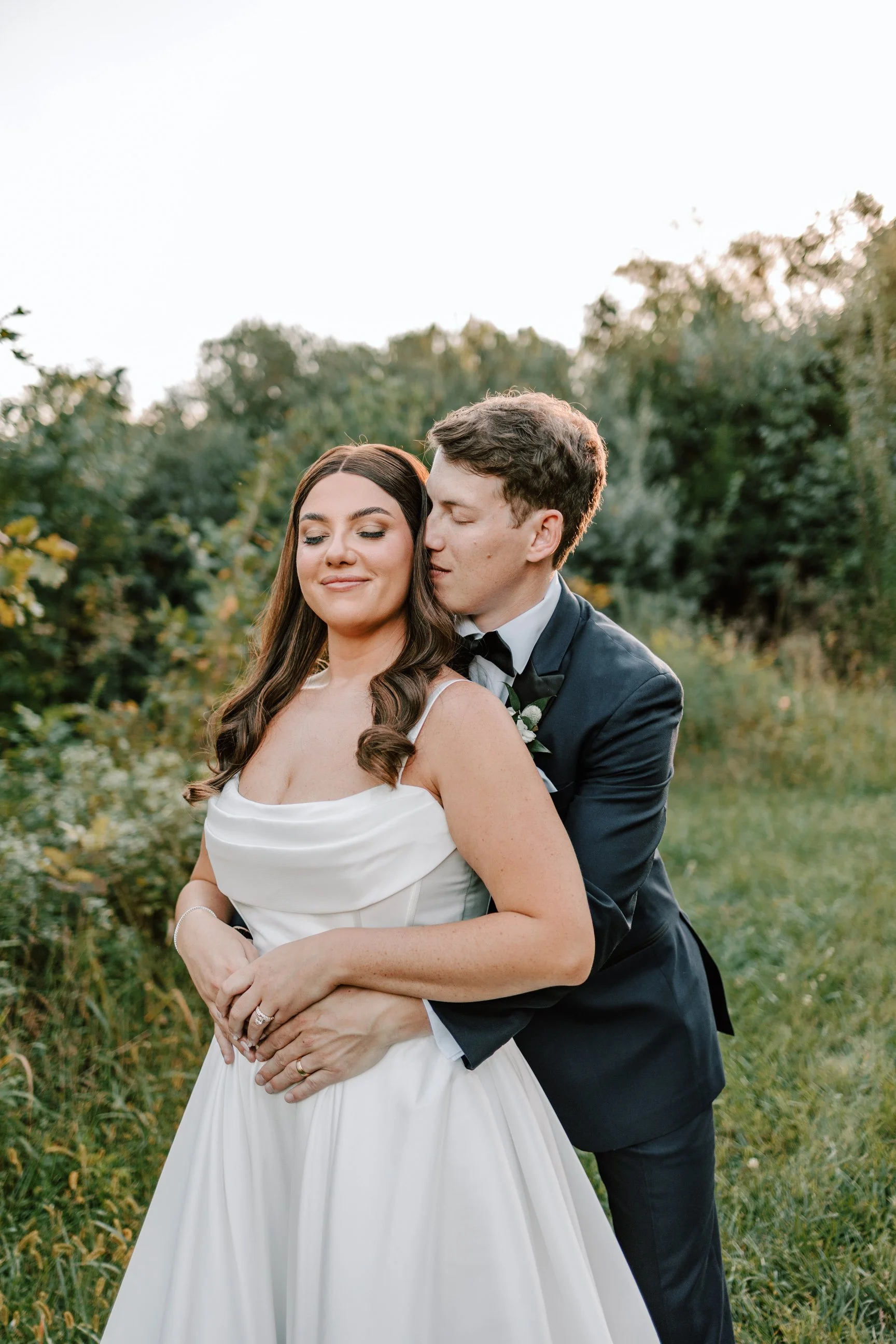 A newlywed couple standing outdoors in a grassy area with trees in the background during sunset, with the groom embracing the bride from behind, both smiling peacefully.