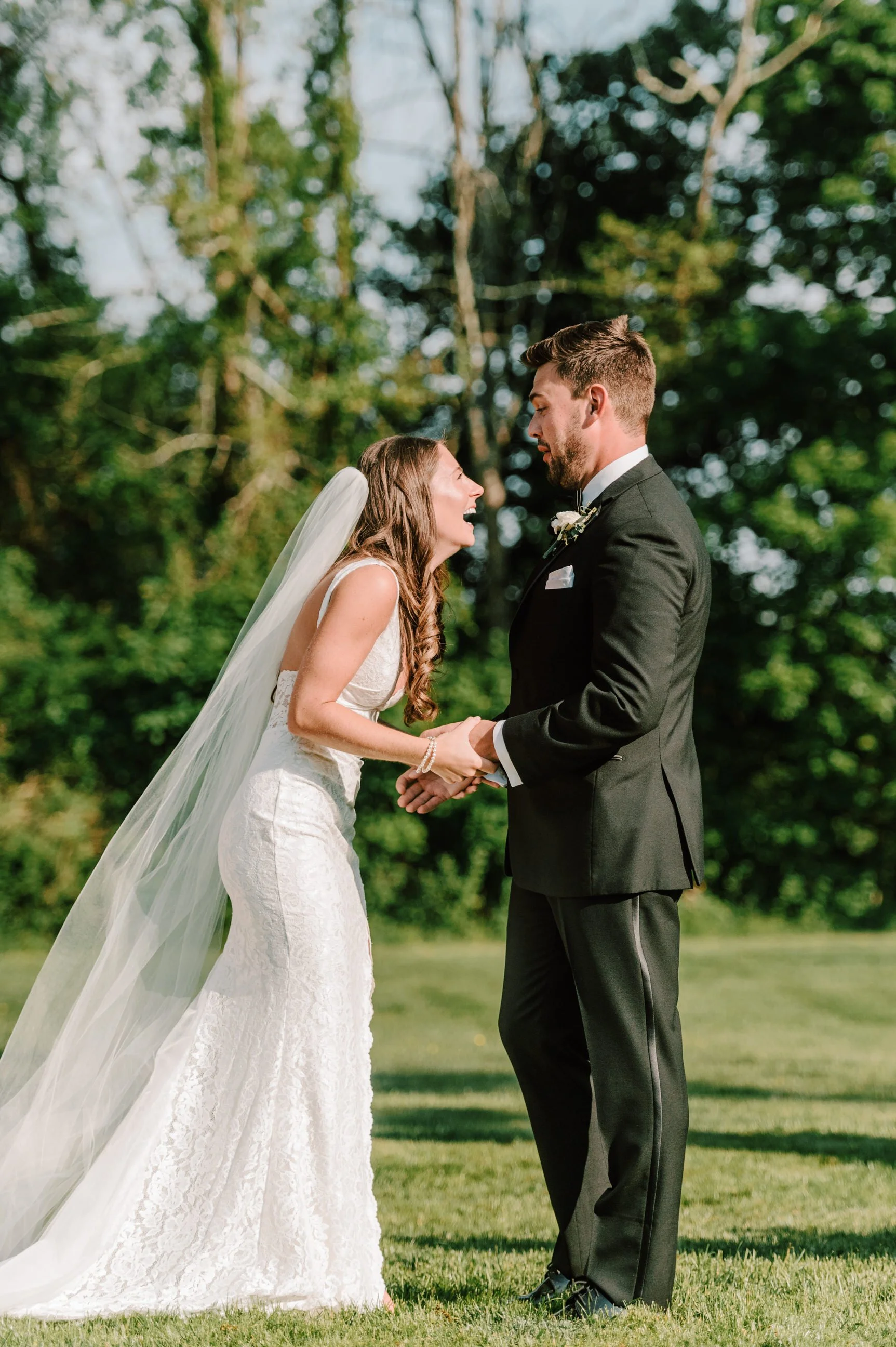 A bride and groom holding hands, smiling, and looking at each other outdoors during their wedding.