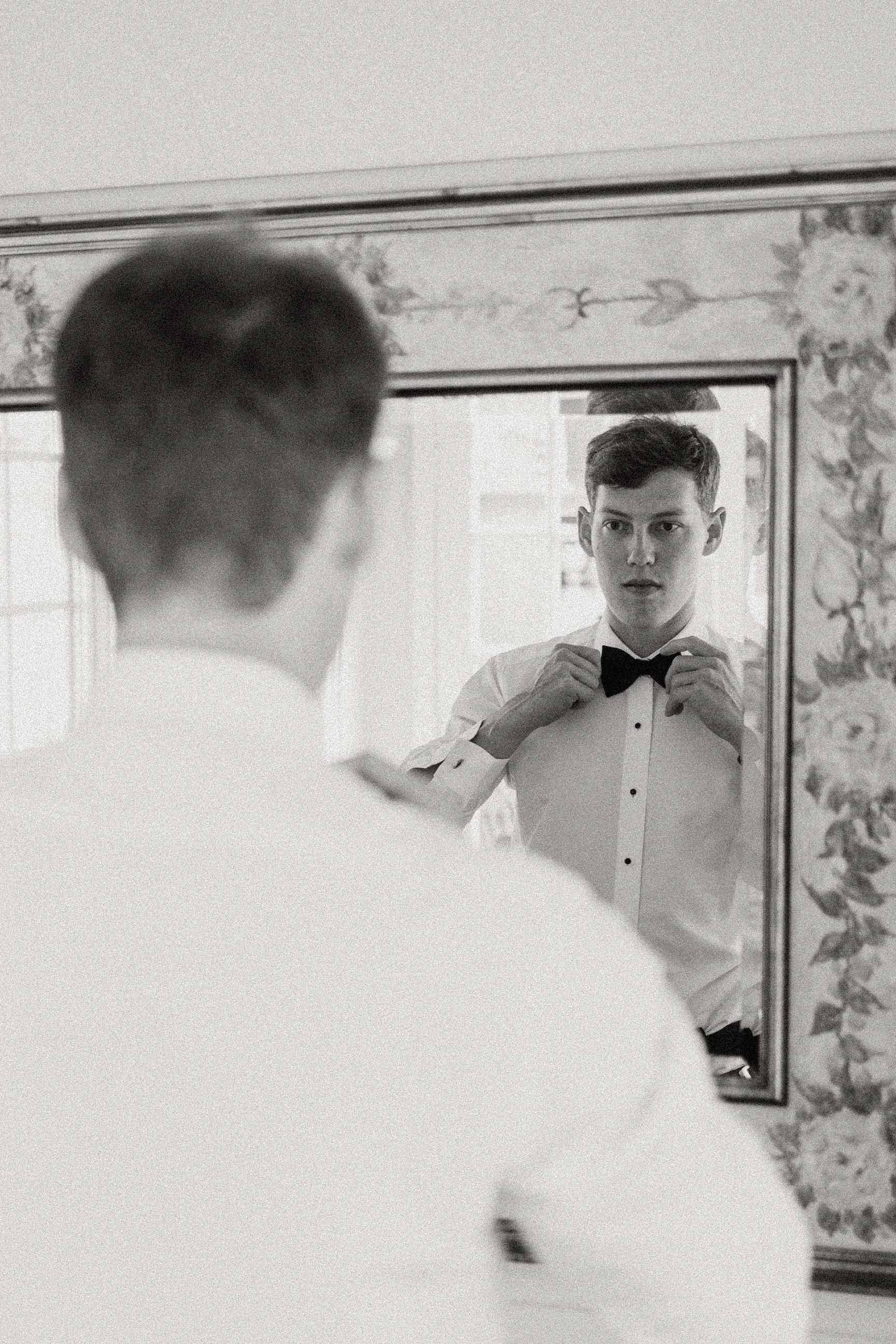A young man adjusts a bow tie while looking at himself in a mirror.