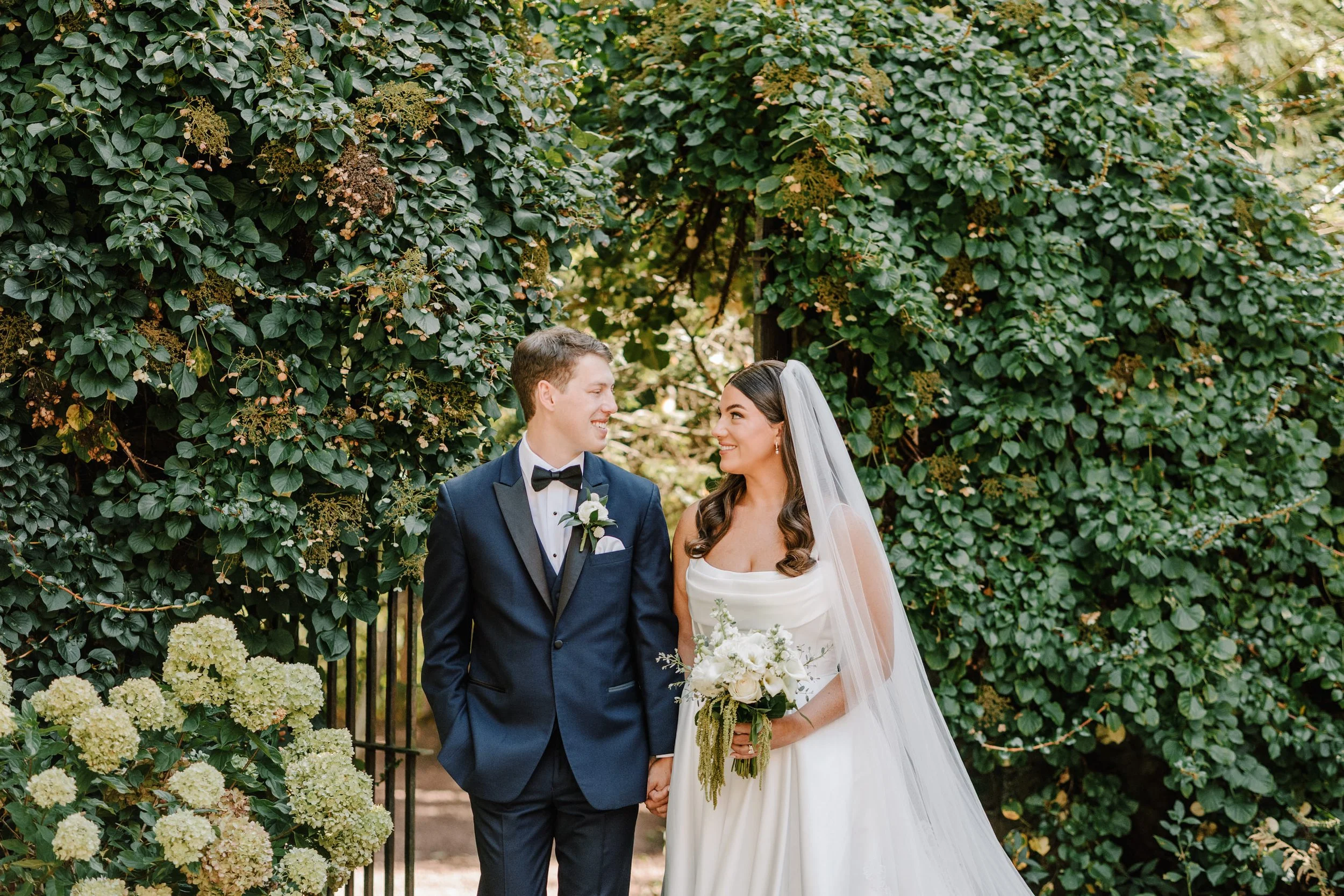 A newlywed couple stands in front of lush green foliage during their wedding photos. The groom is wearing a navy tuxedo with a black bow tie and boutonnière, while the bride is in a white wedding gown holding a bouquet of white flowers. They are smil