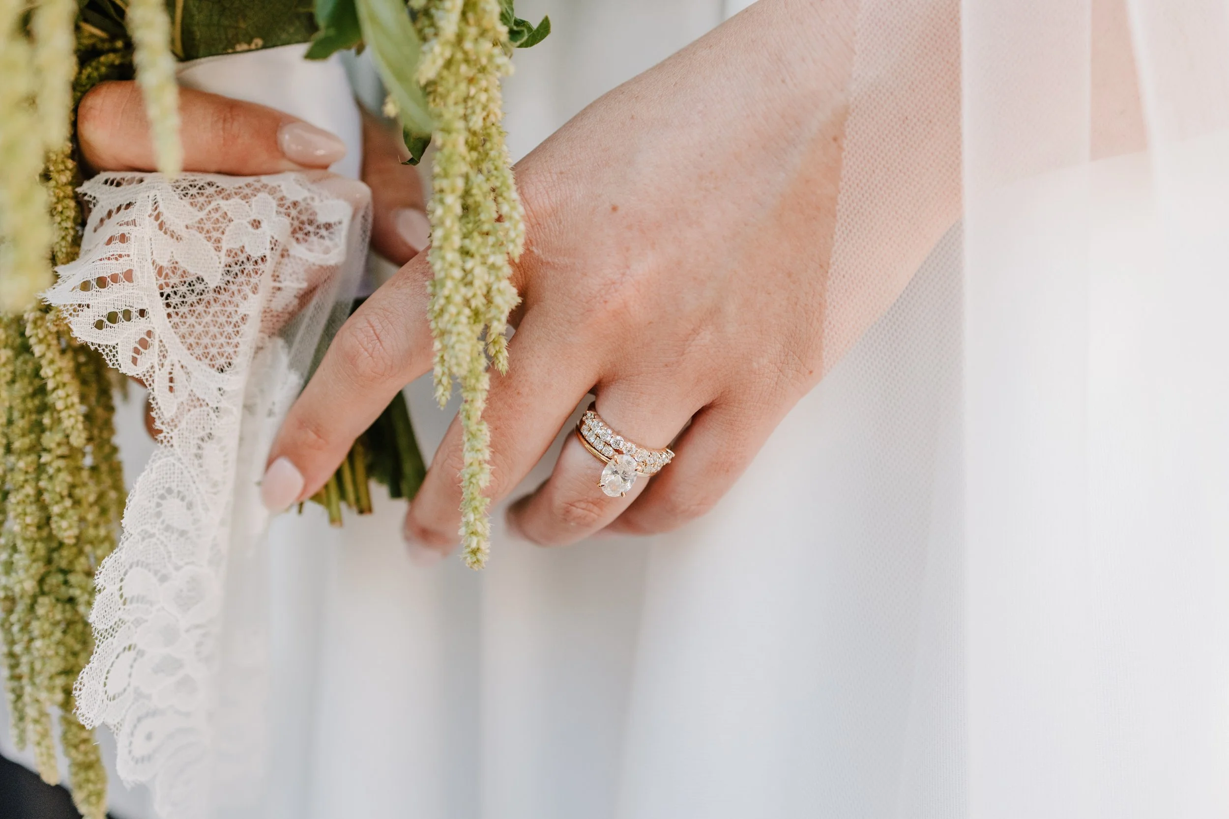 Close-up of a woman's hand with a wedding ring and engagement ring, holding a bouquet with lace and greenery.
