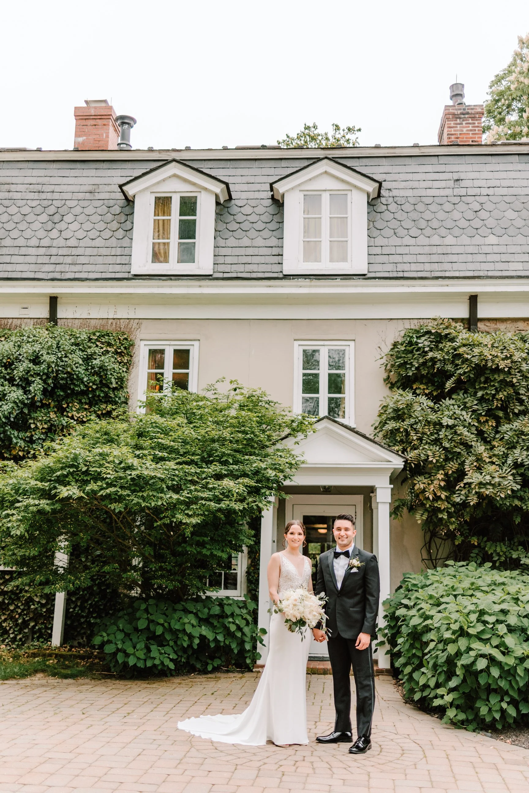 A bride and groom standing outside a house on a wedding day, holding hands, with greenery and a brick walkway in front.