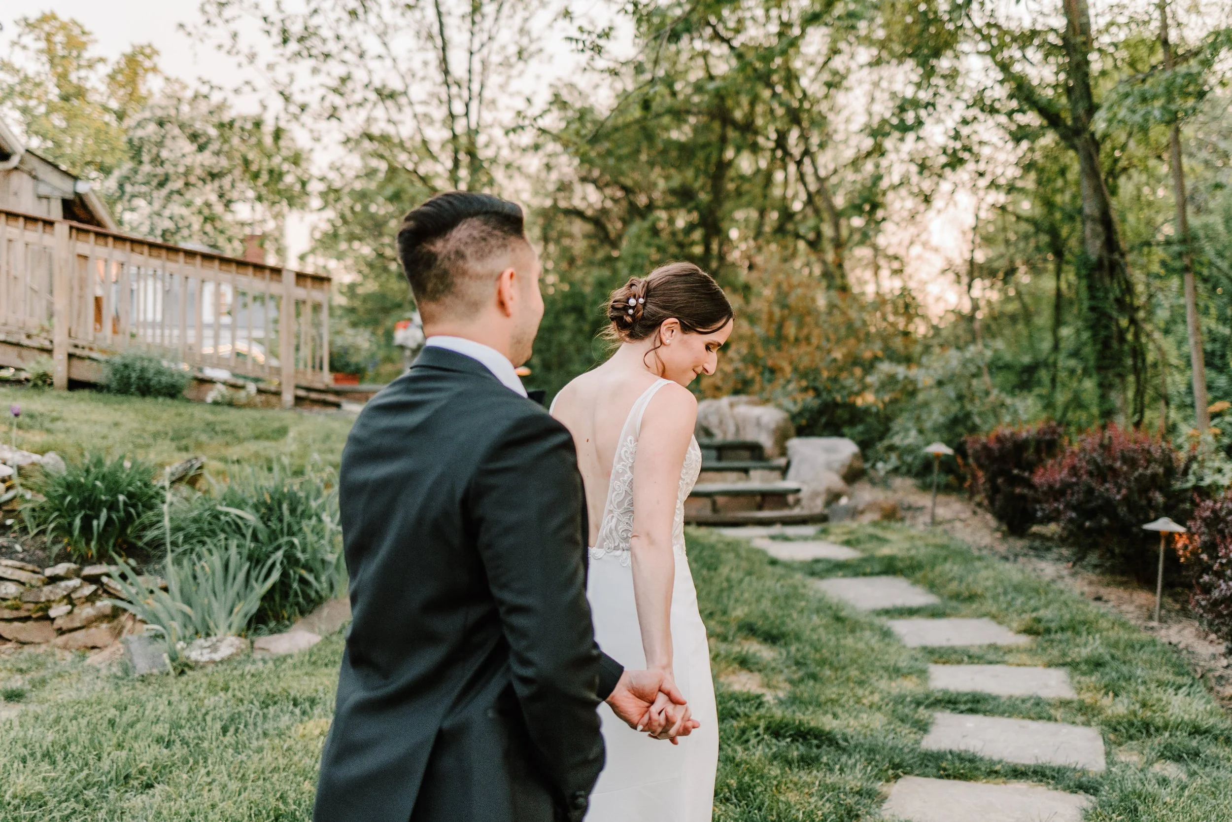 A bride and groom holding hands outdoors, walking on a stone path in a lush garden with trees and bushes, during sunset.