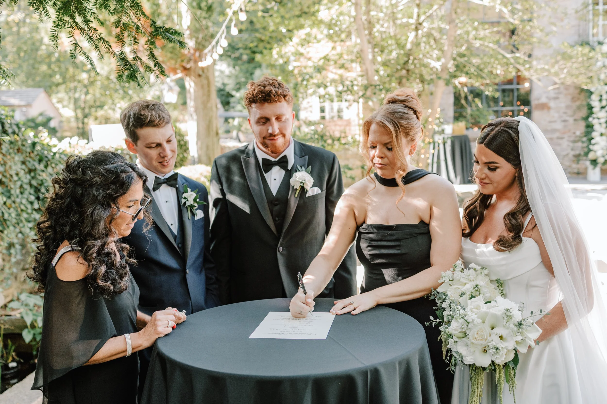 A bride and groom signing a marriage certificate during their outdoor wedding ceremony, surrounded by bridesmaids and groomsmen, in a garden setting with trees and string lights.