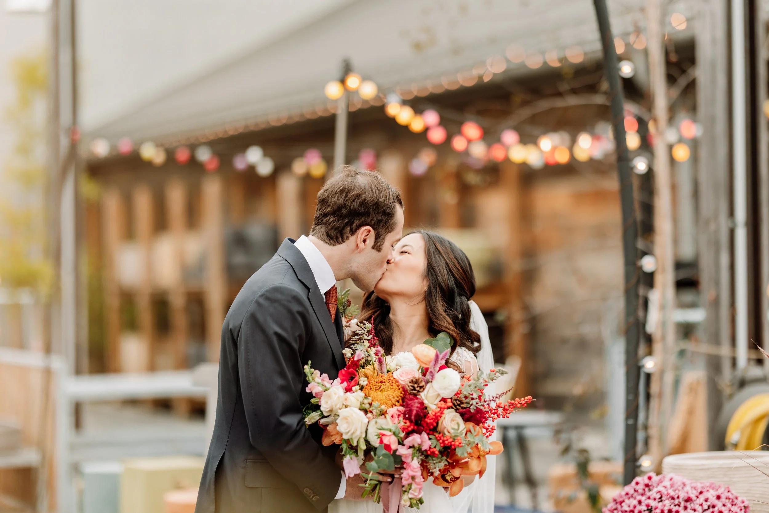 A newlywed couple sharing a kiss at their wedding reception, with the bride holding a large bouquet of flowers. The background features string lights and a rustic outdoor setting.
