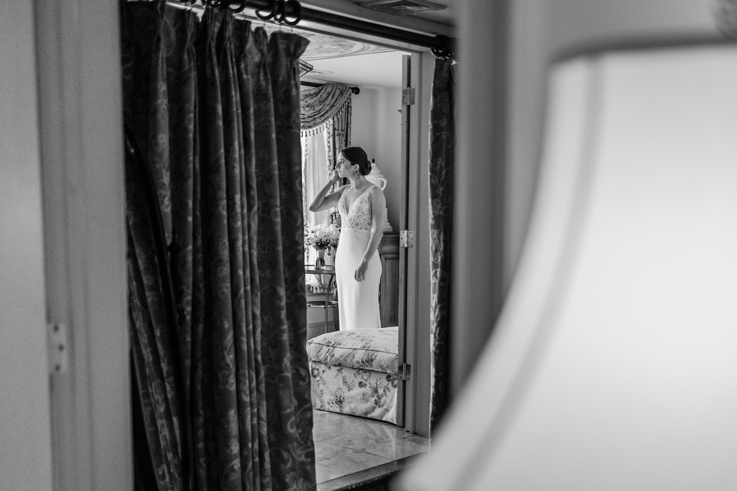 A woman in a wedding dress looking at herself in a mirror, seen through a partially open door with curtains.