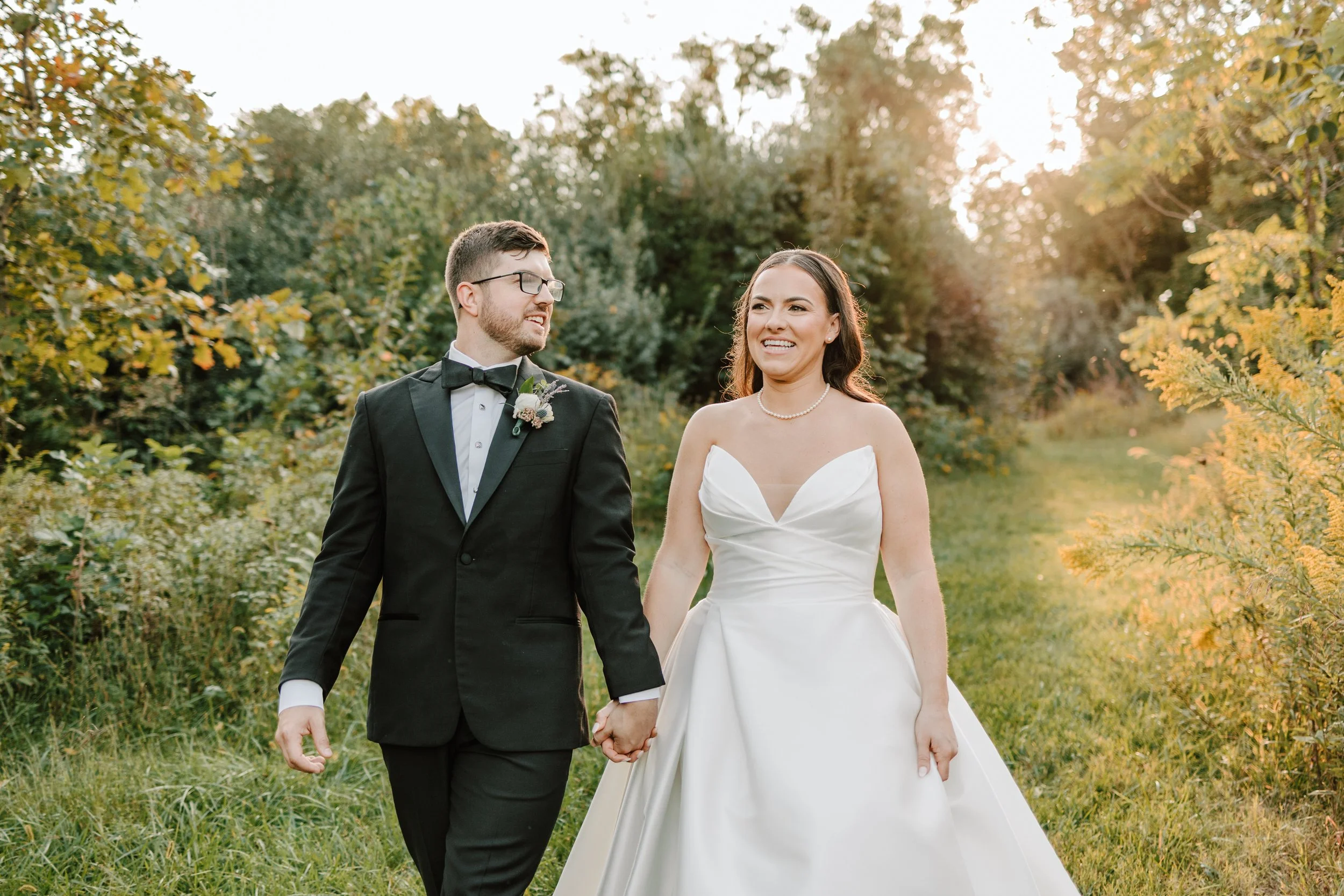 A newlywed couple walking hand in hand outdoors on a grassy path during sunset, surrounded by lush greenery and yellow-orange foliage.