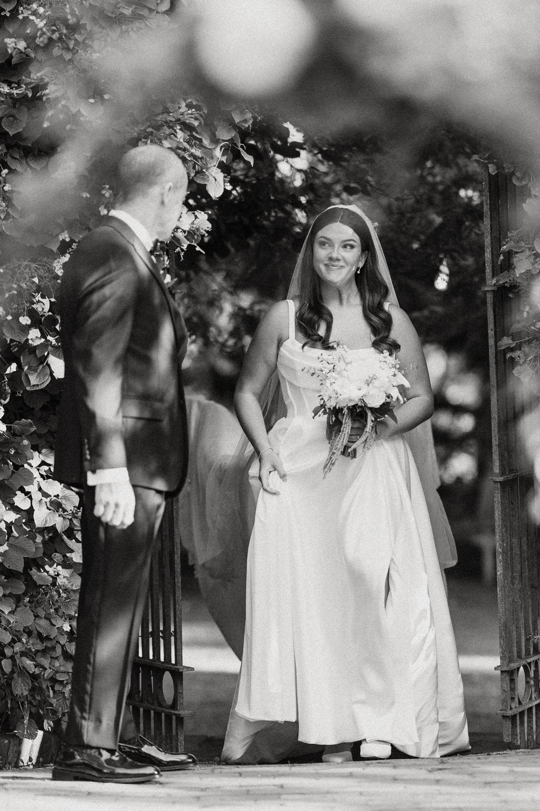 Black and white photo of a bride in a wedding dress holding a bouquet, smiling at a groom in a tuxedo, standing in a garden with trees and an open gate.