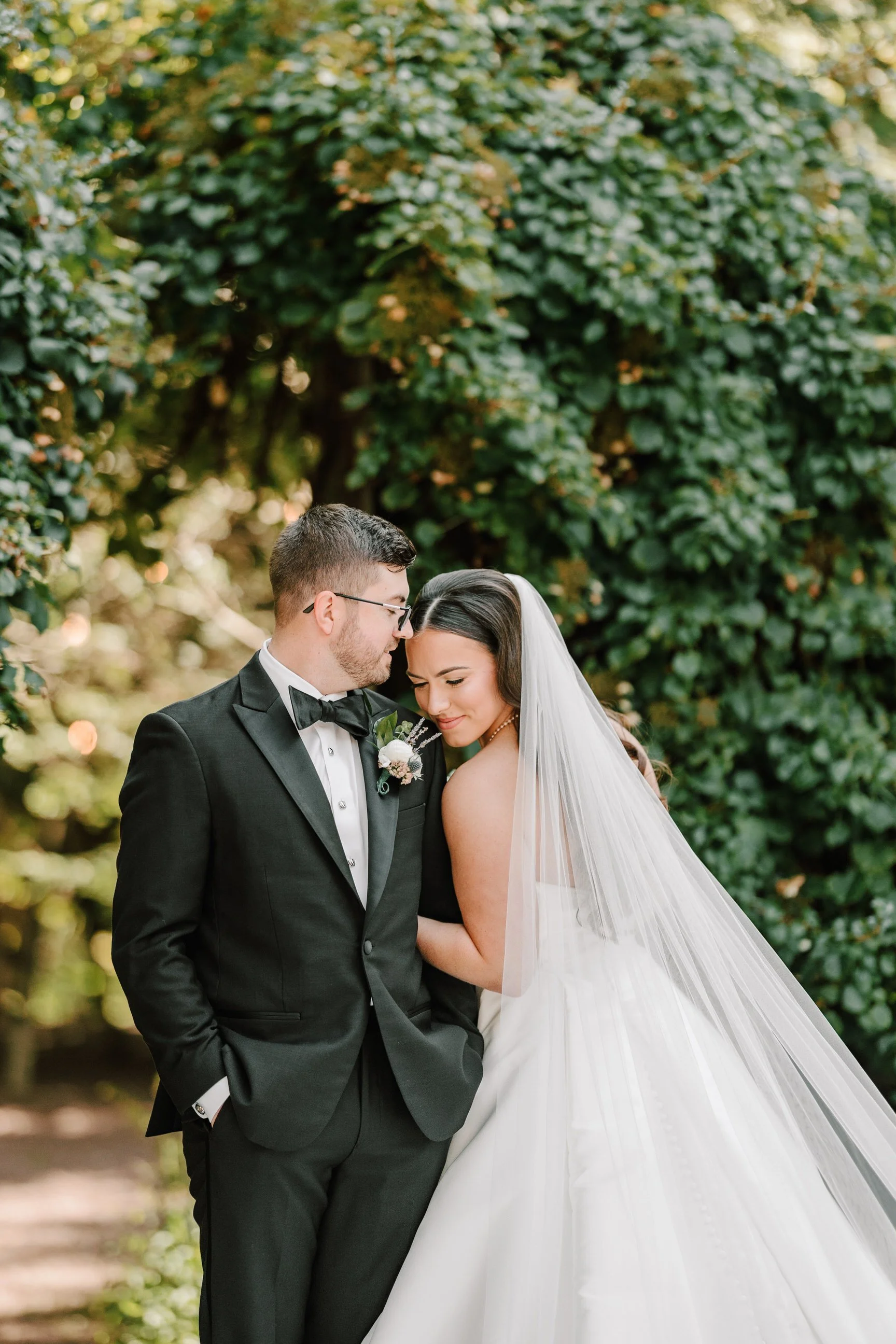 A bride and groom stand close together outdoors surrounded by lush greenery, with the couple sharing an intimate and tender moment at their wedding. Philadelphia wedding photographer