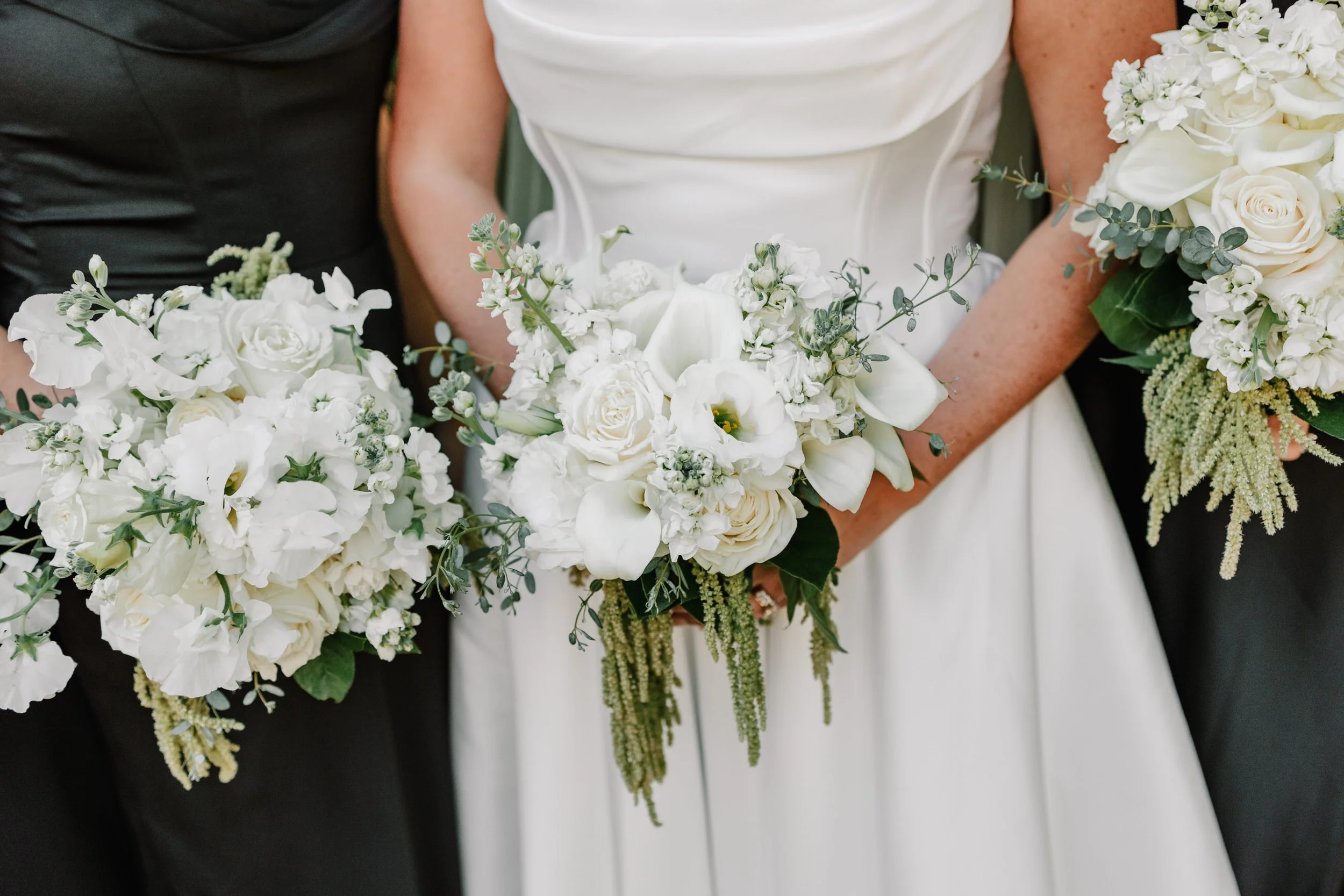 Close-up of a bride holding a white flower bouquet at a wedding. The bride is wearing a white dress, and near her, there are two other people holding similar bouquets, one in black attire.