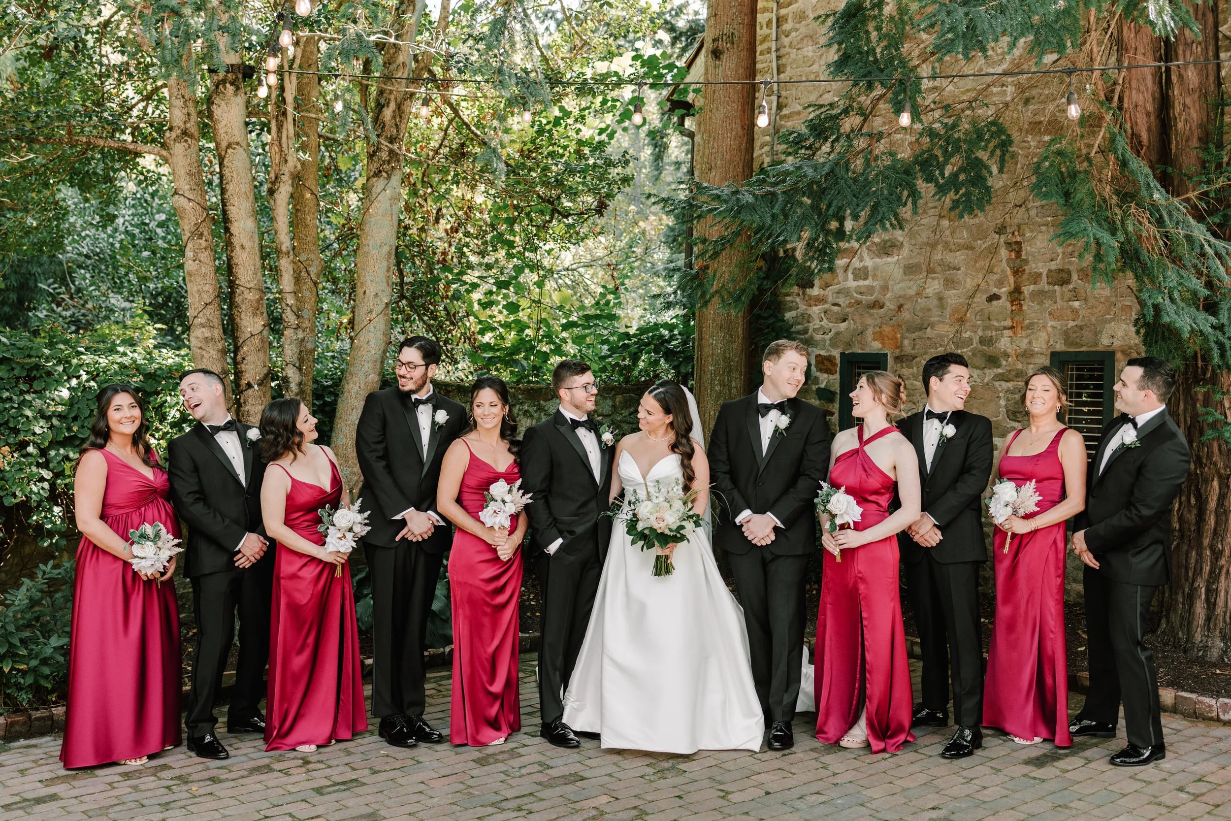 Wedding party of twelve people, six women in red dresses holding bouquets and six men in black tuxedos, outdoors with trees and stone building in the background.