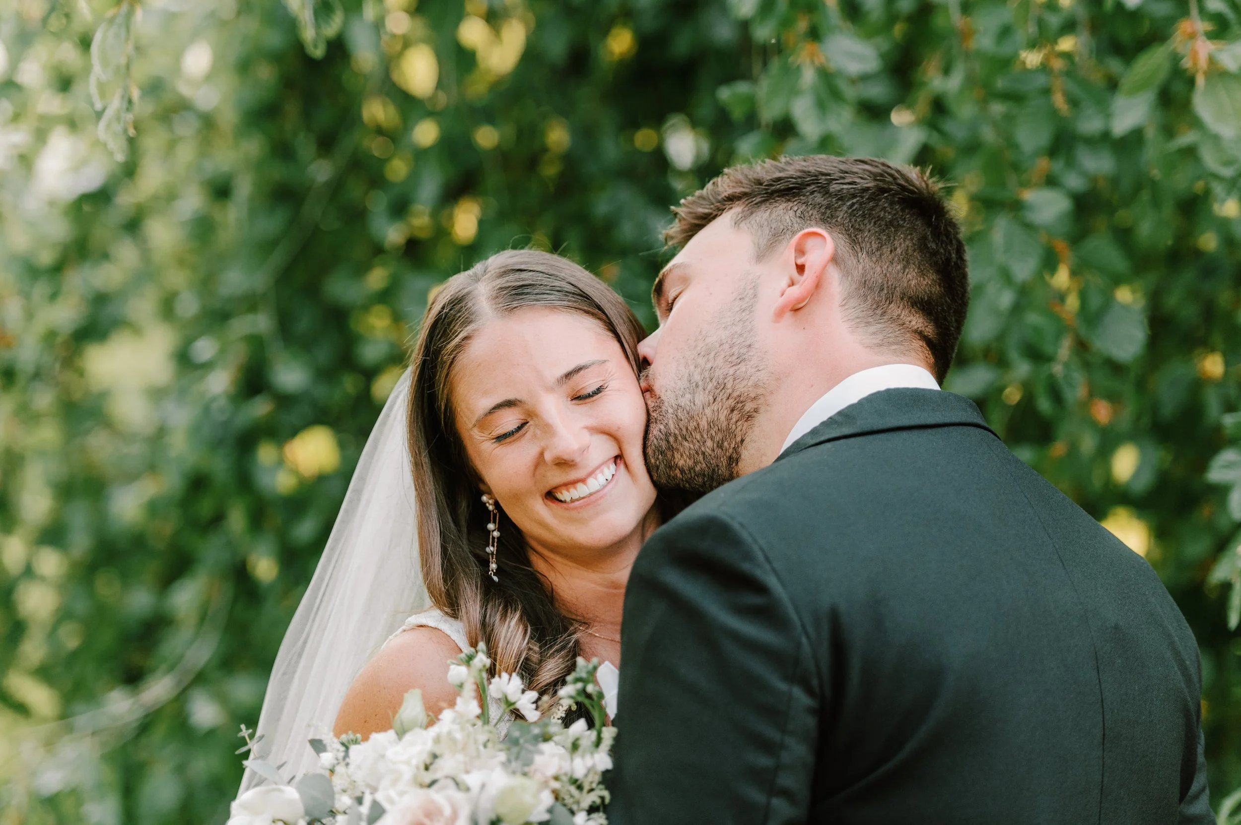 A bride and groom sharing a kiss at their wedding outdoors, with the groom kissing the bride on her cheek, and a background of green foliage.