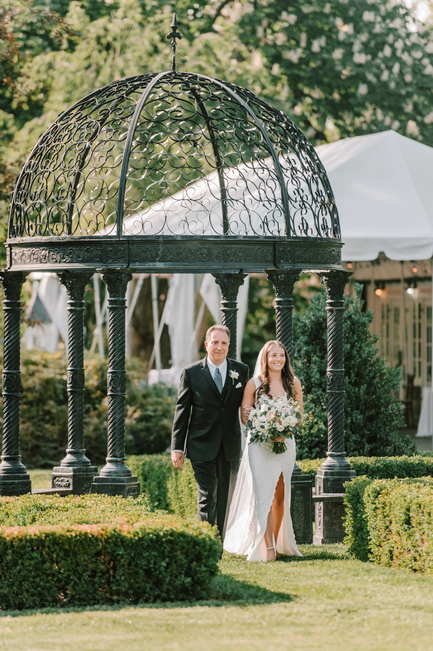 A bride in a white wedding dress holding a bouquet walks down the aisle with a man in a black suit, beneath a decorative black metal gazebo in a garden setting.