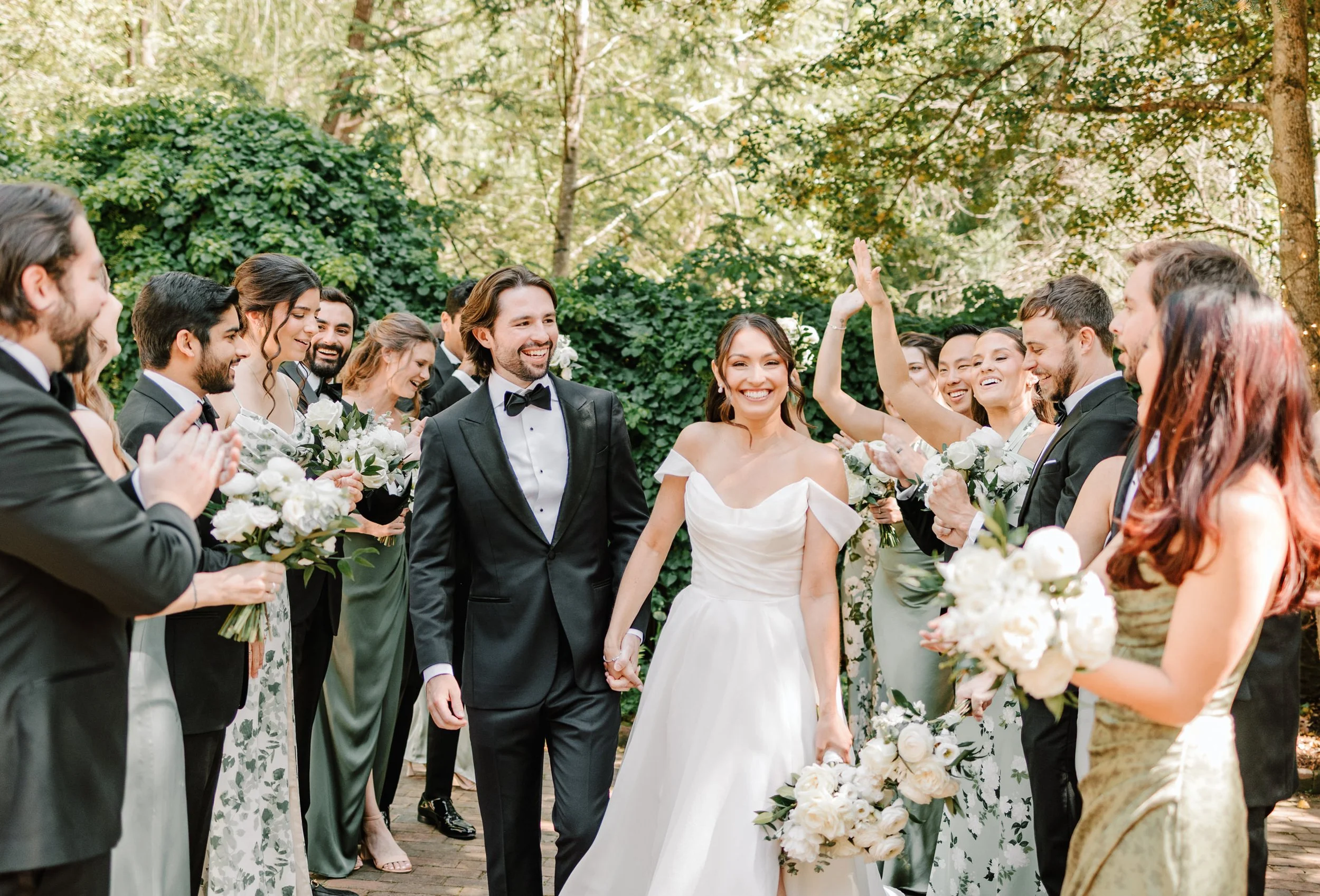 A bride and groom walking hand in hand with a wedding party celebrating outdoors, surrounded by friends in formal attire, with lush green trees in the background.