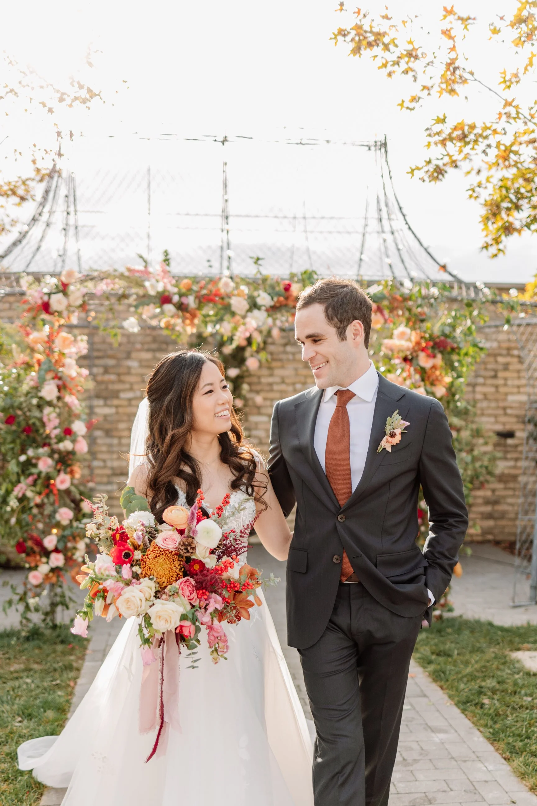 A bride and groom smiling at each other during their wedding ceremony outdoors, with a floral arch and brick wall in the background.