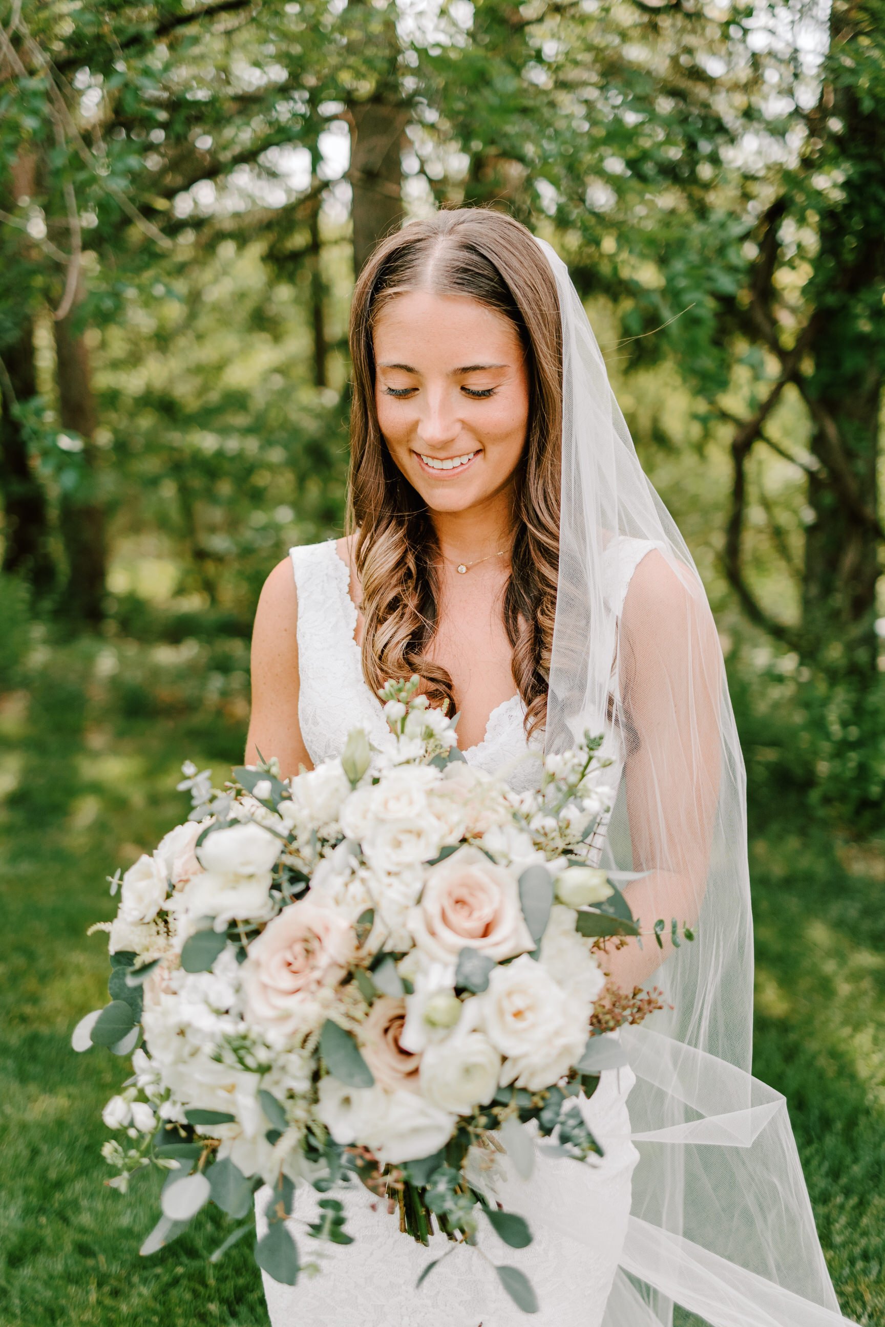 A smiling bride in a white wedding dress holding a large bouquet of white and blush roses with greenery, standing outdoors with green trees in the background.