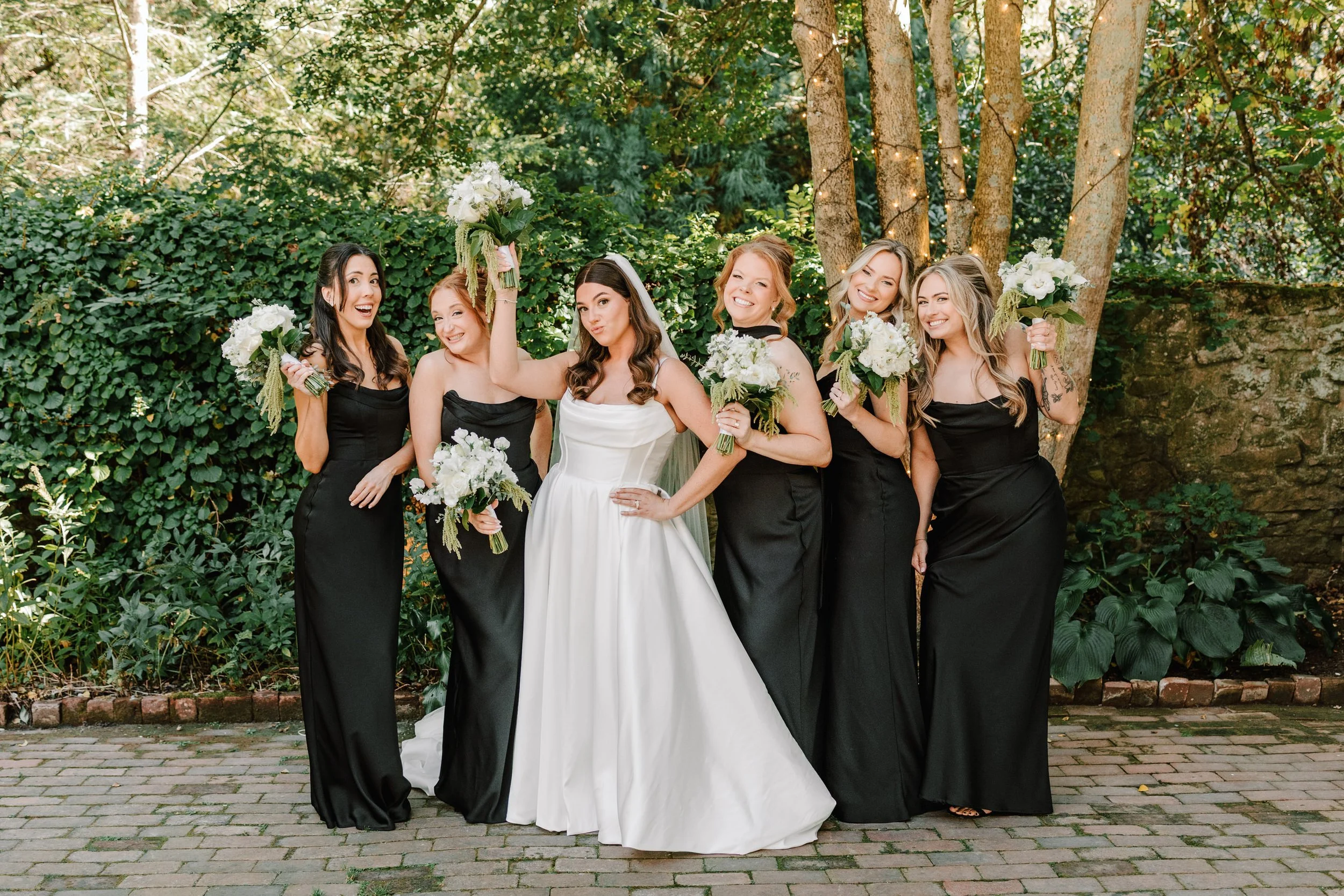 A bride in a white wedding gown with dark brown hair in loose curls stands with her five bridesmaids dressed in black, holding bouquets of white flowers, outdoors with lush greenery and trees in the background.