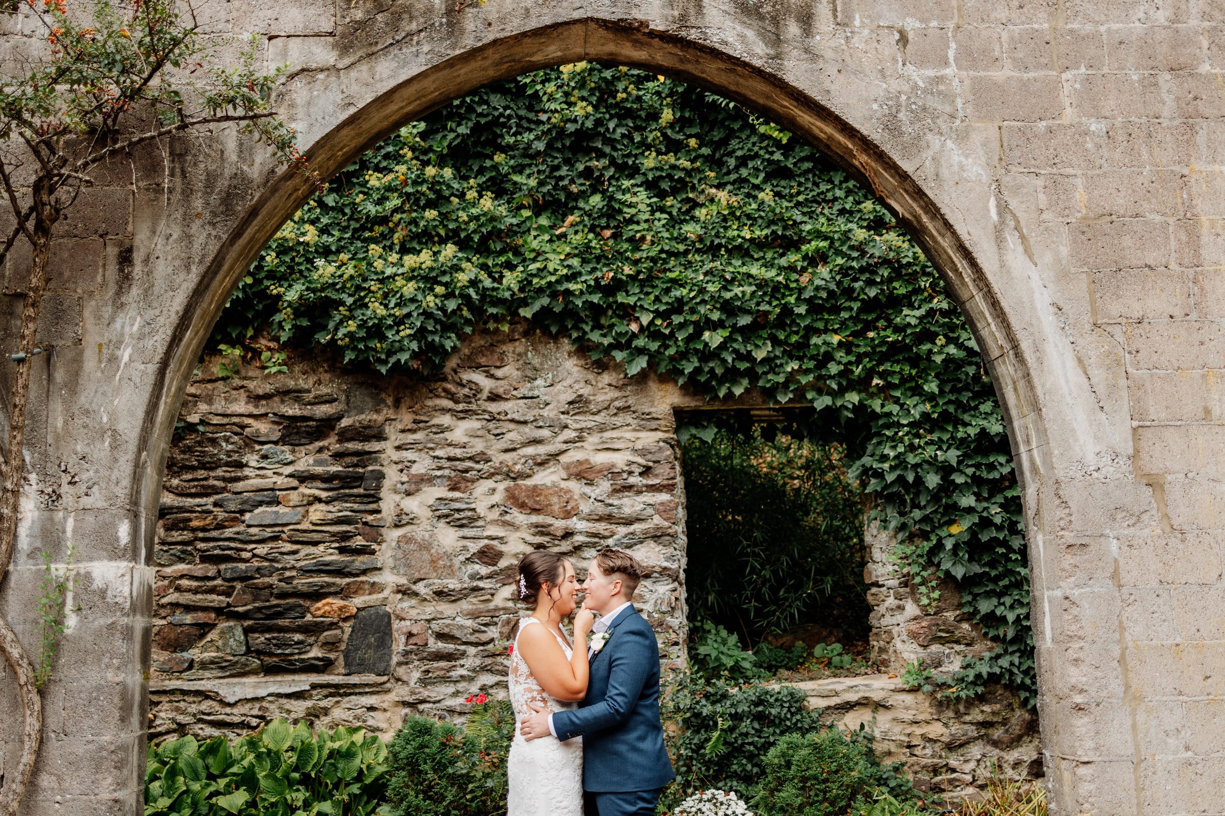 A couple in wedding attire shares an intimate moment under a stone archway covered in ivy, surrounded by lush greenery and an old brick wall.