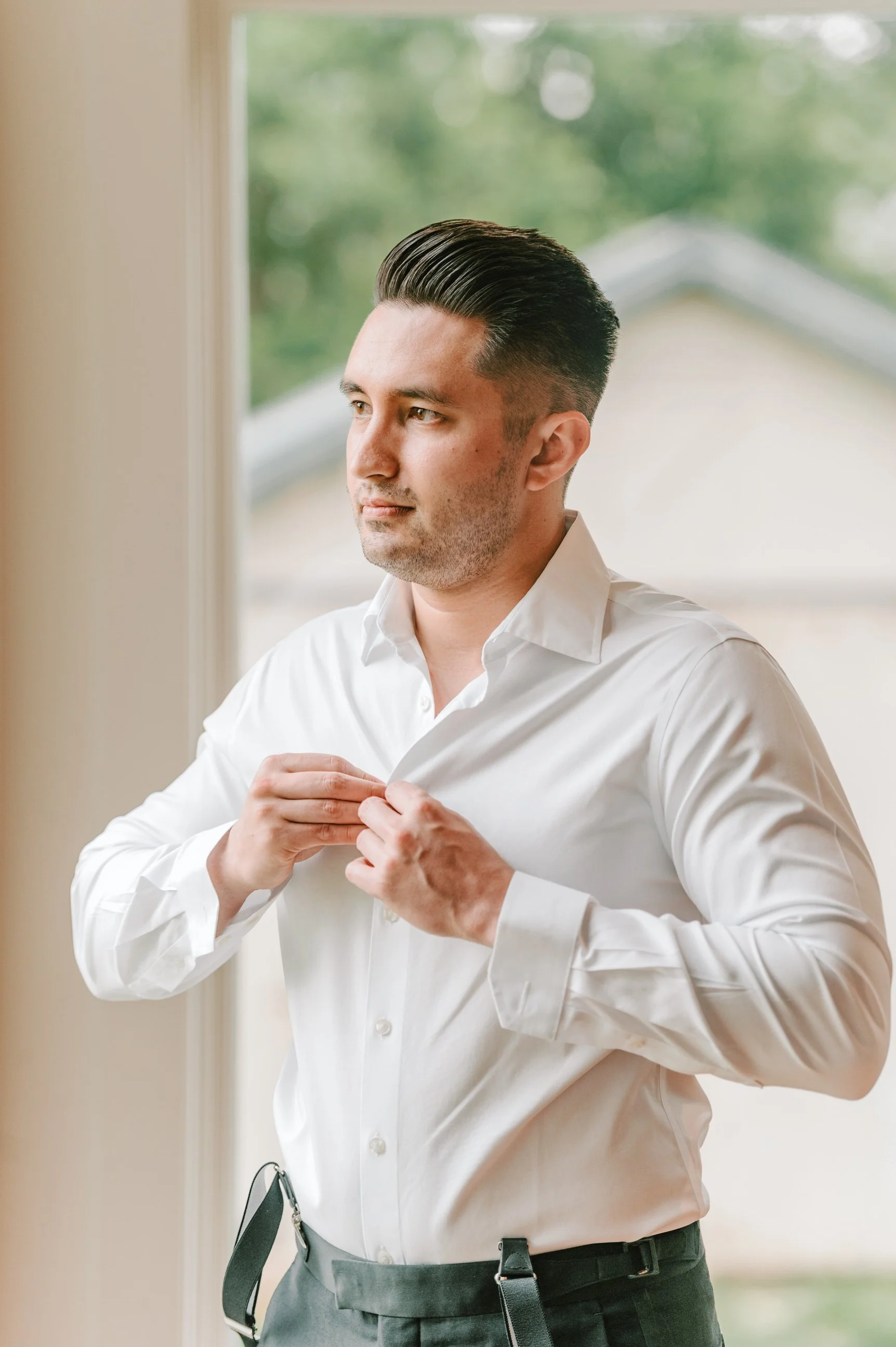 A man in a white dress shirt buttoning his shirt near a window with a blurred outdoor background.