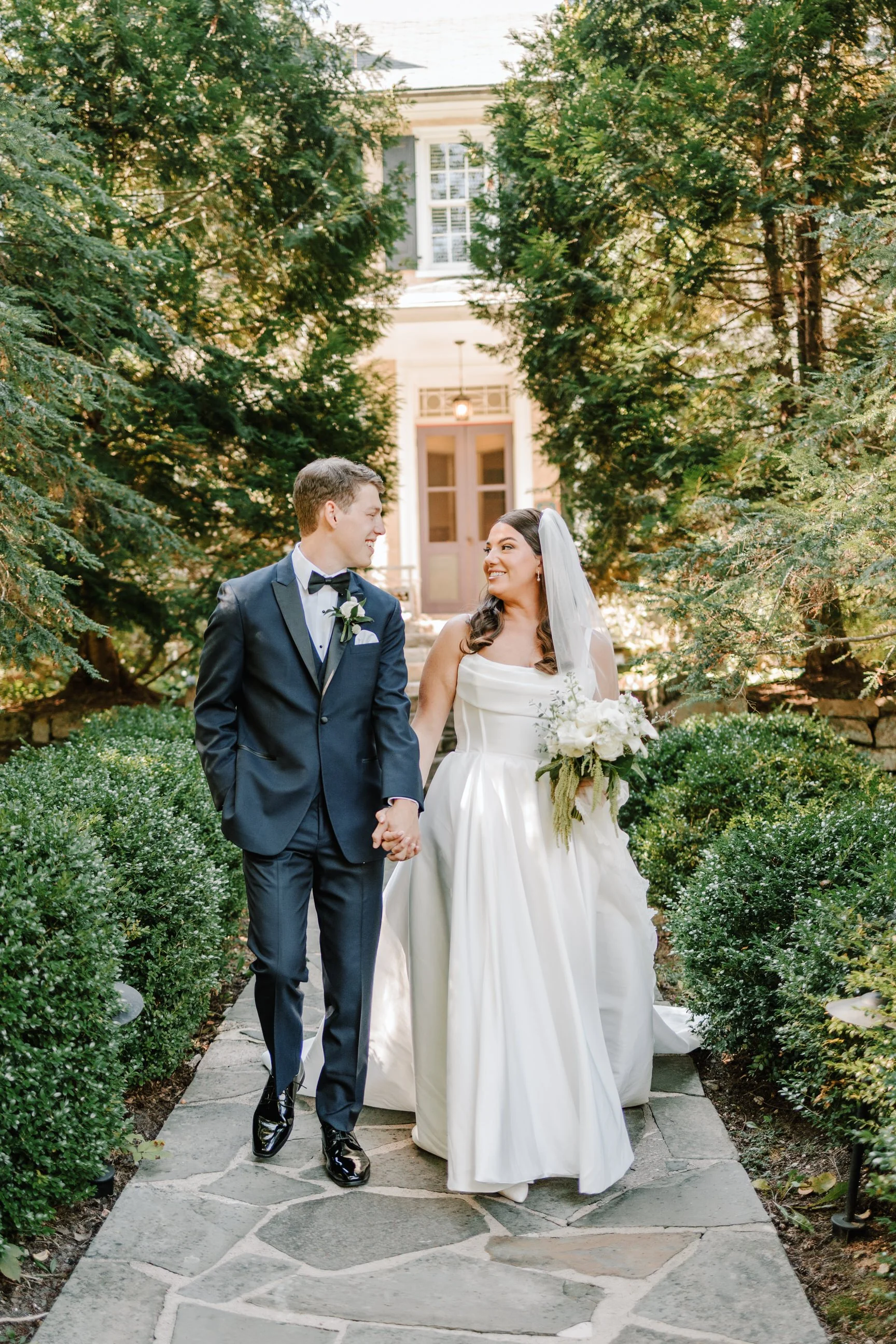 A bride and groom holding hands and smiling at each other while walking on a stone pathway in a garden with green shrubs and tall trees, in front of a large house.