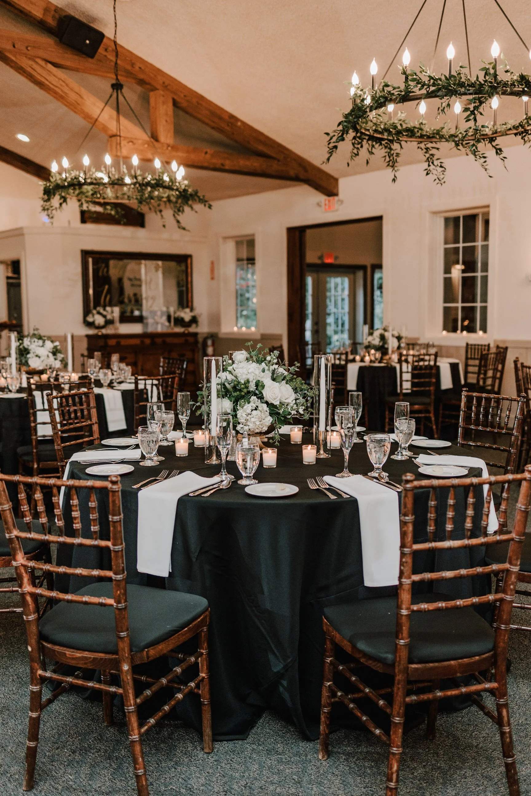 A beautifully decorated banquet hall with a round table set for a formal event. The table features a floral centerpiece with white flowers and greenery, candles, and glassware. The chairs are wooden with dark cushions. The room has wooden beams and h