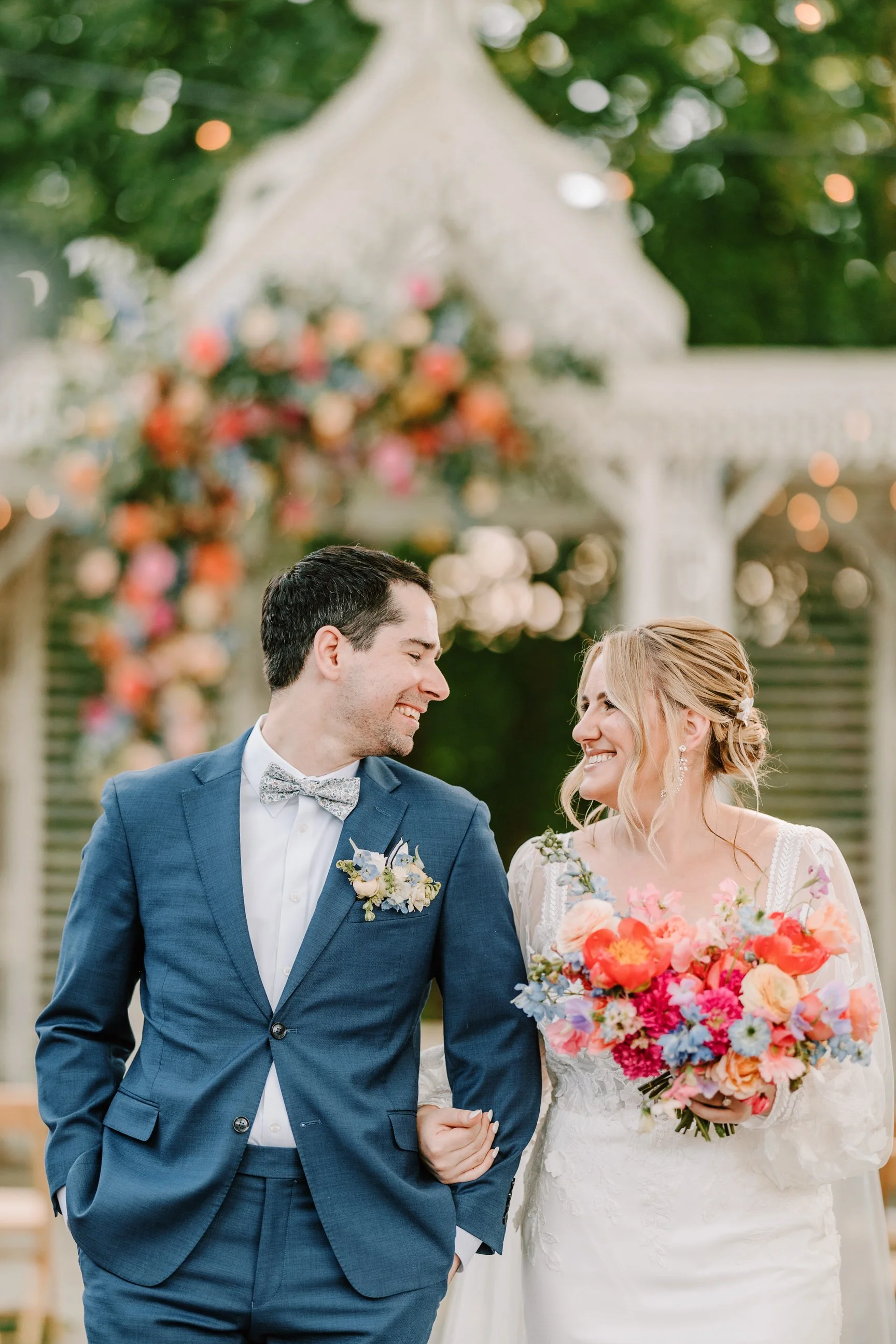 Bride and groom smiling at each other; bride holding colorful bouquet; outdoor wedding setting with floral arch.