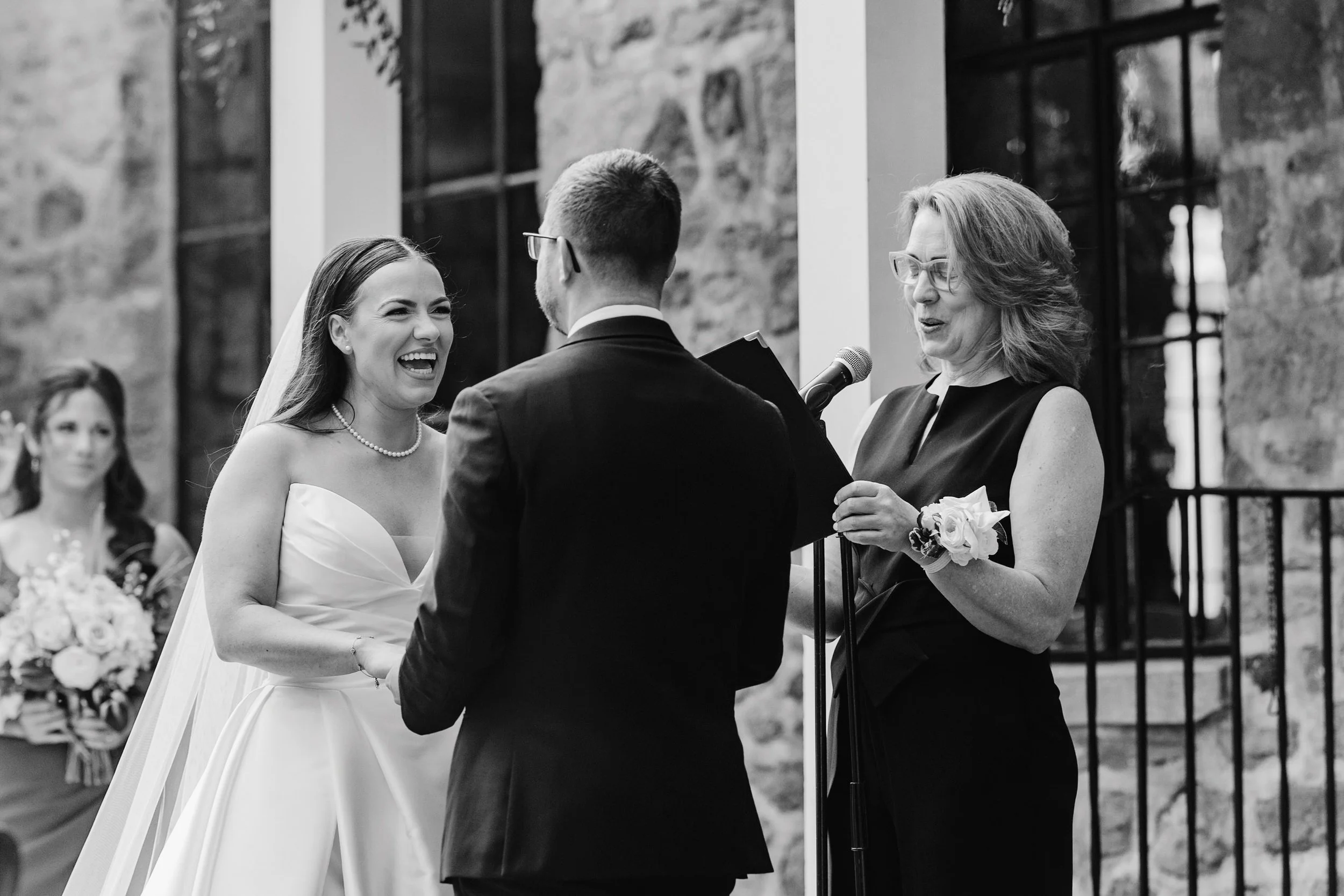 Black and white photo of a bride laughing during a wedding ceremony, standing in front of an officiant reading vows, with a groom facing her. A bridesmaid holding a bouquet is in the background.