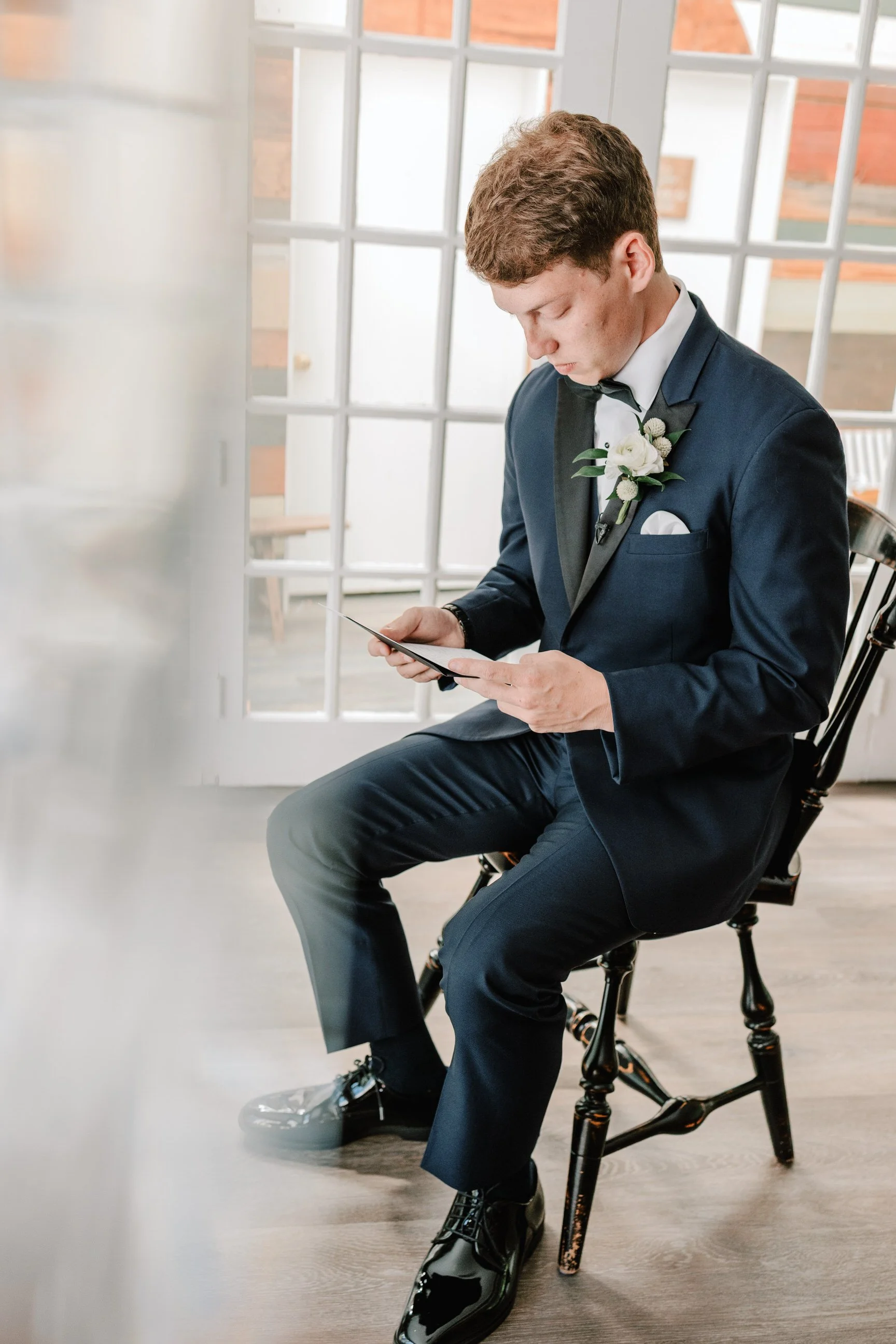 Young man in a navy tuxedo with a white boutonniere sitting on a black wooden chair, looking at a small black notebook or card, indoors near large glass-paned doors.