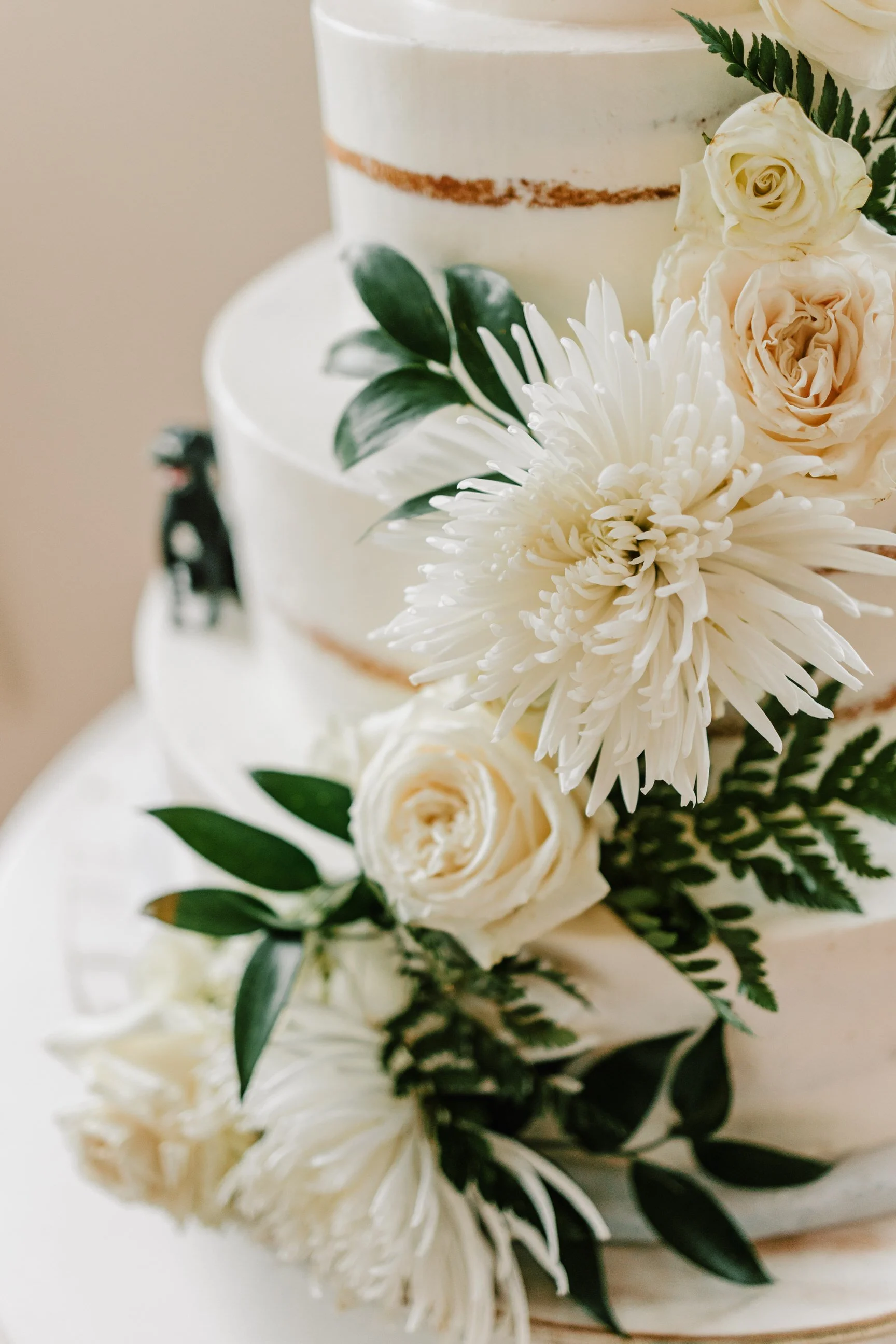 Close-up of a multi-tiered white wedding cake decorated with white roses, a large white chrysanthemum, green leaves, and fern fronds.