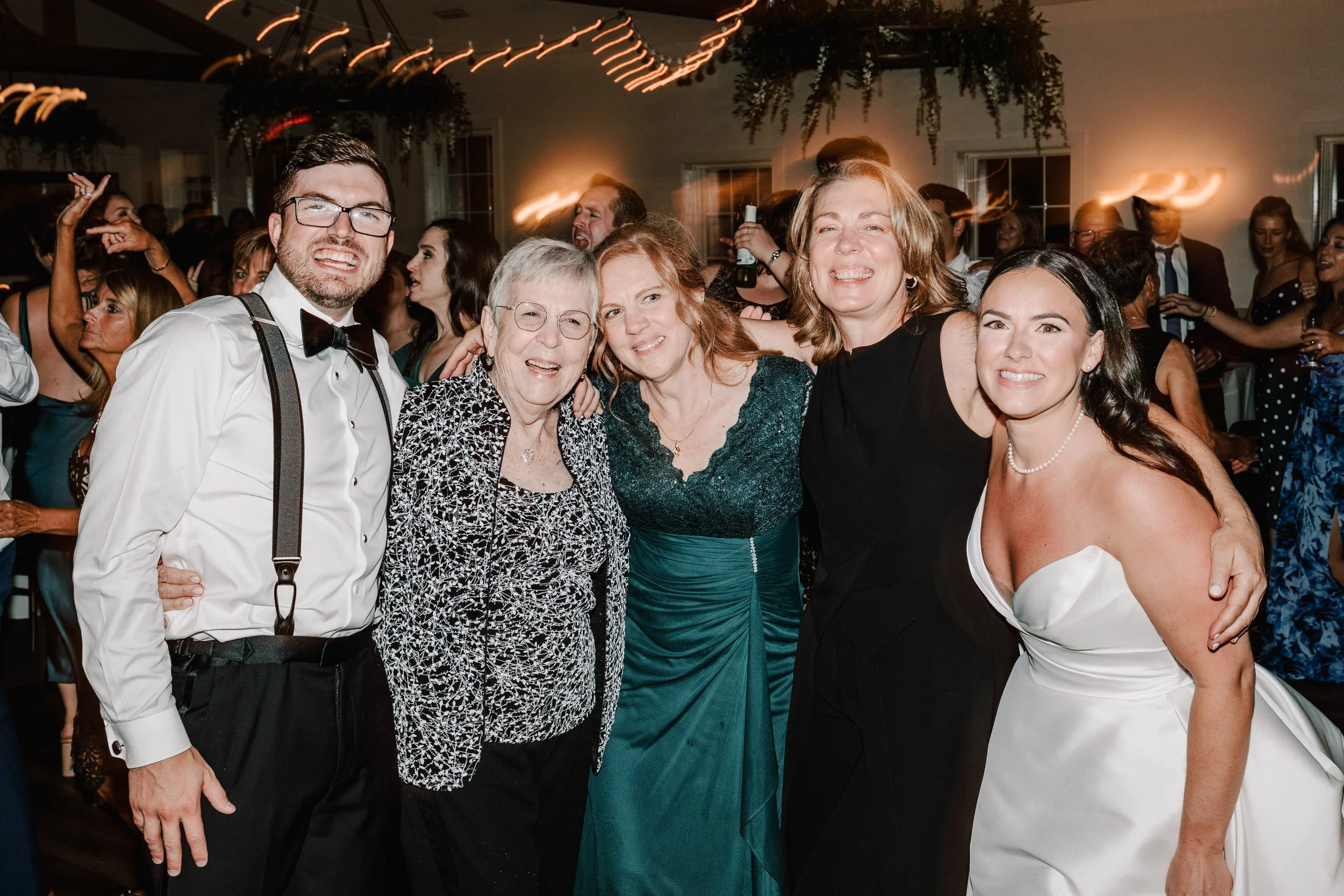 Group of five people smiling and posing together at a celebration or wedding reception, with dancing and lively crowd in the background.