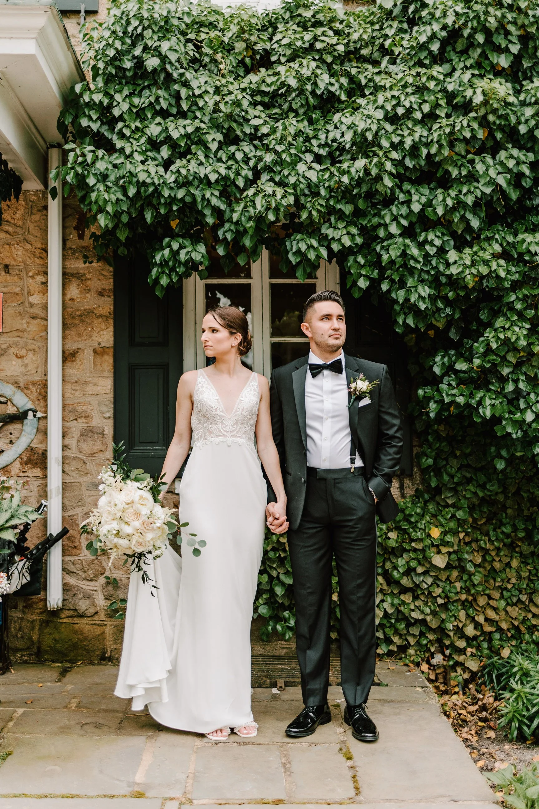 A bride and groom holding hands stand outdoors in front of a stone building with ivy-covered wall and a window. The bride wears a white lace wedding gown and holds a bouquet, while the groom wears a black tuxedo with a bow tie and a boutonniere.
